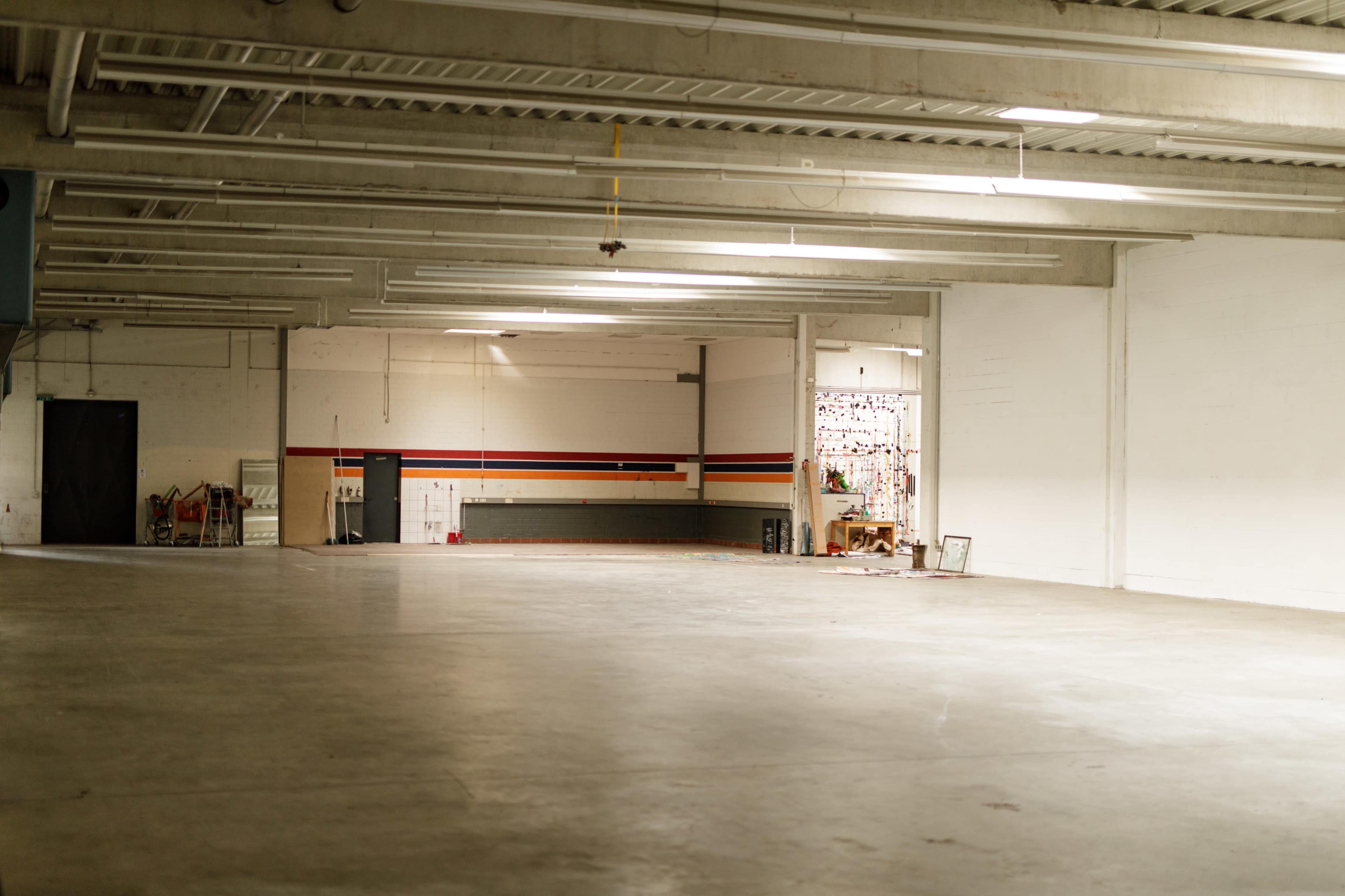 The image shows a large, empty warehouse space with concrete flooring and exposed ceiling beams, featuring a partially visible area with shelves and items against one wall.