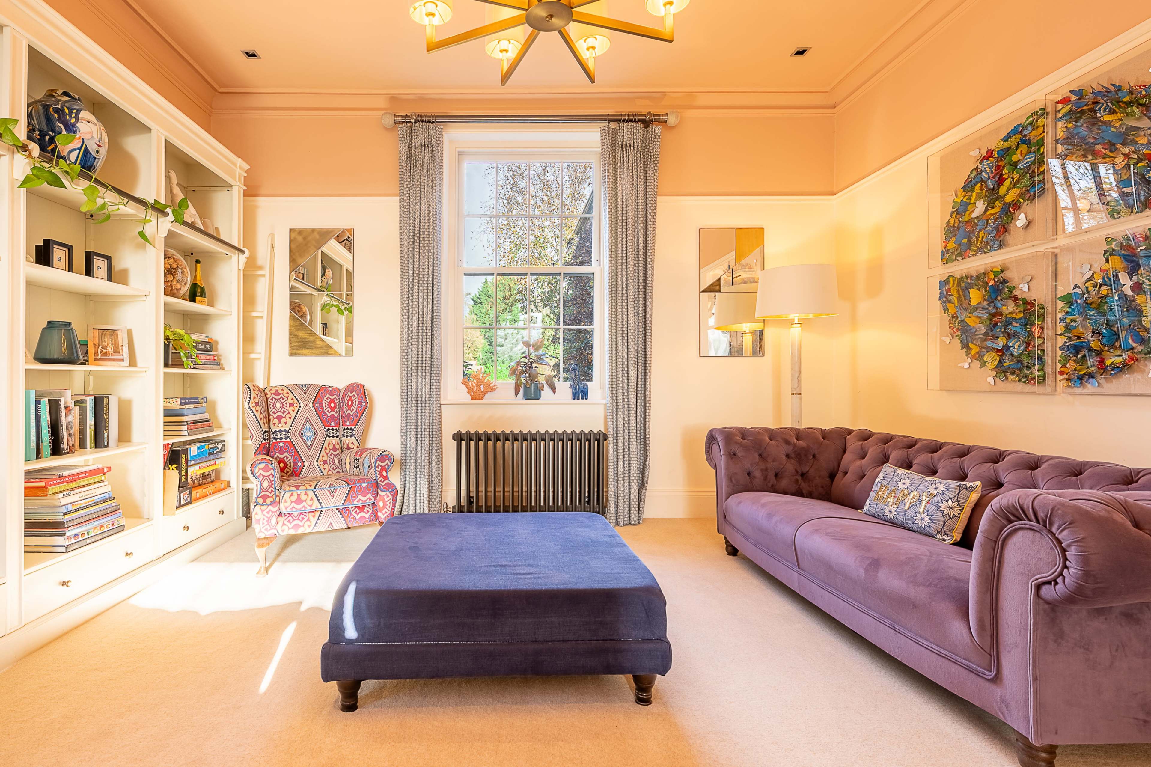 A brightly lit living room features a purple velvet ottoman, a tufted purple sofa, and a colorful armchair, with bookshelves and a large window showcasing greenery outside.