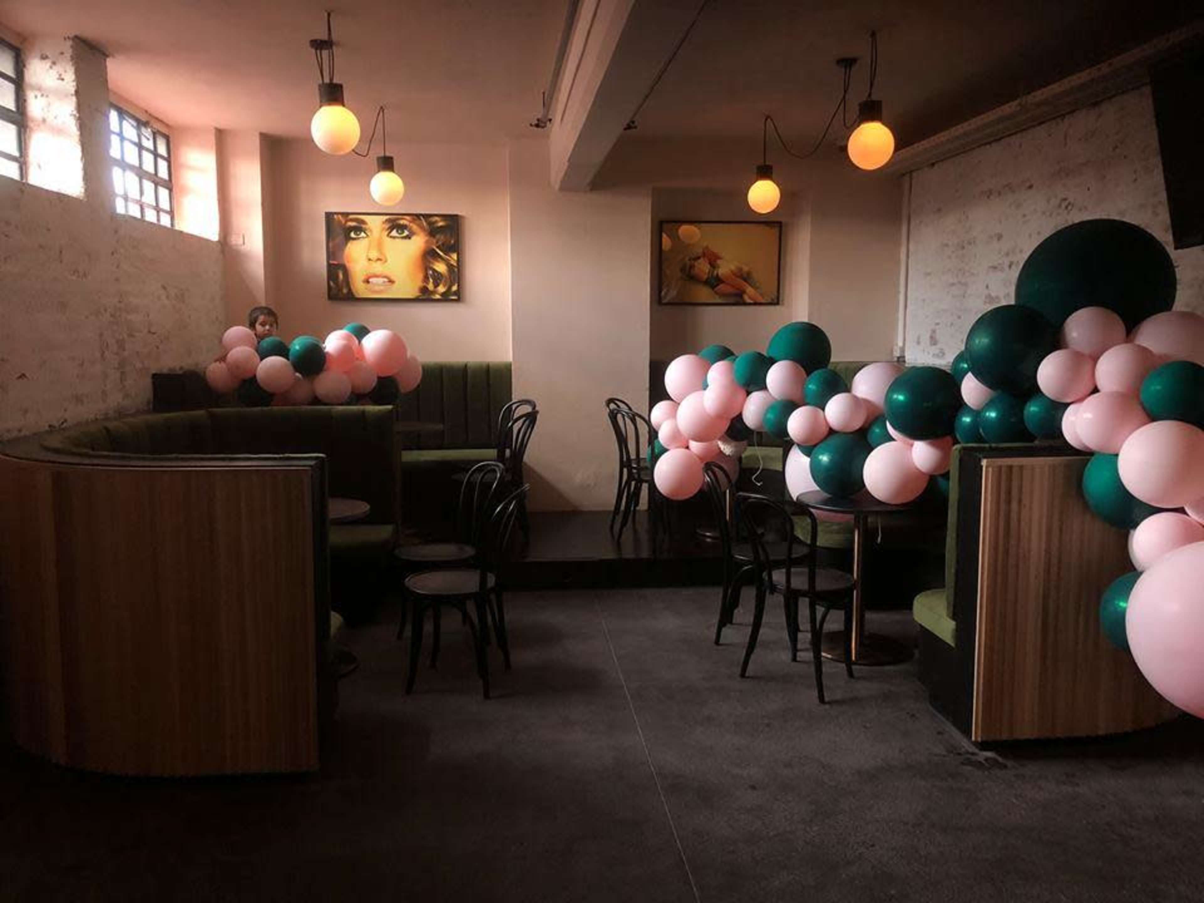 A dimly lit café with green booths, wooden seating, and clusters of pink and green balloons arranged decoratively around the space.