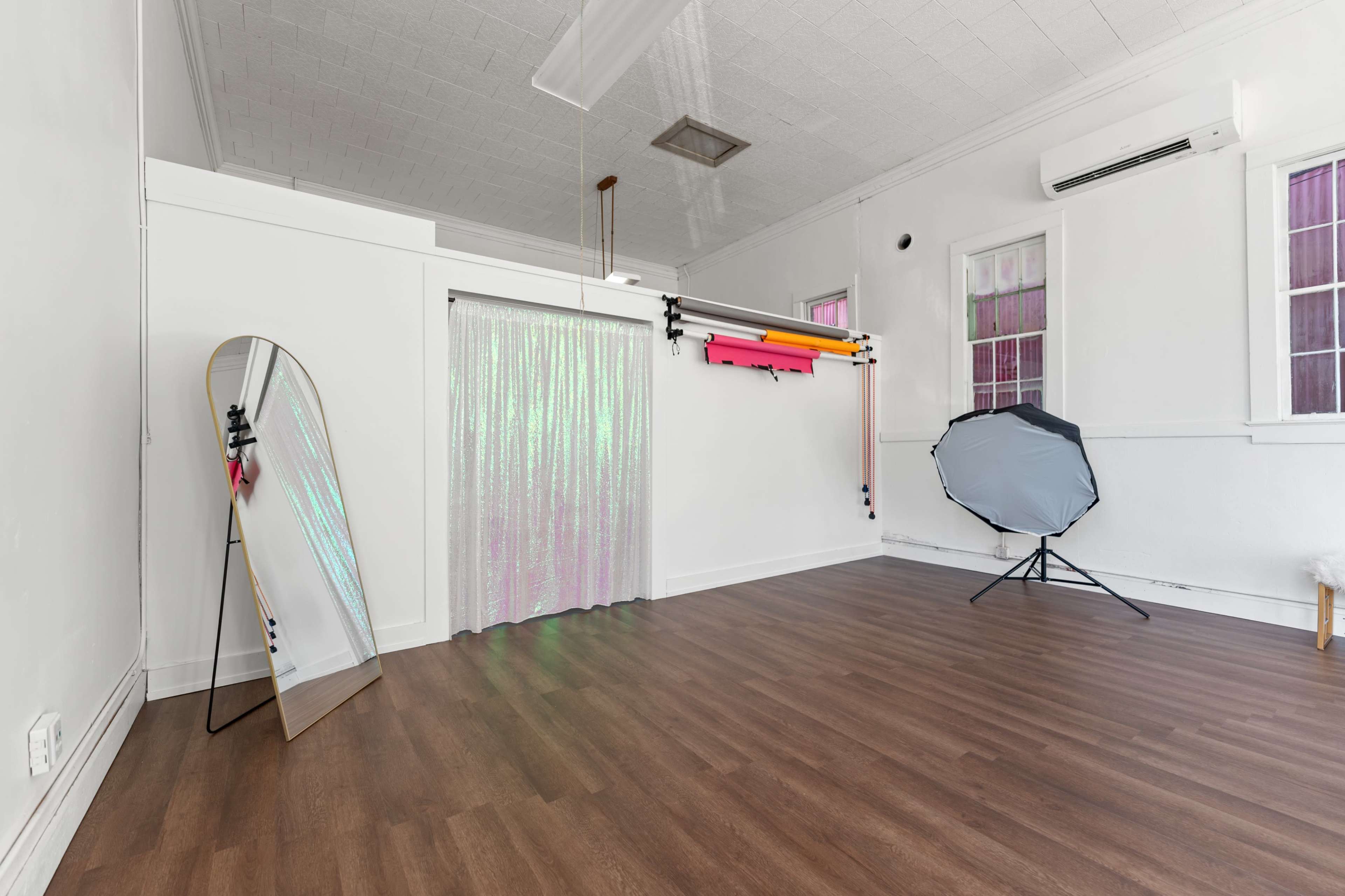 The image shows a spacious, well-lit room with a wooden floor, a large mirror against one wall, and various photography equipment arranged on another wall.