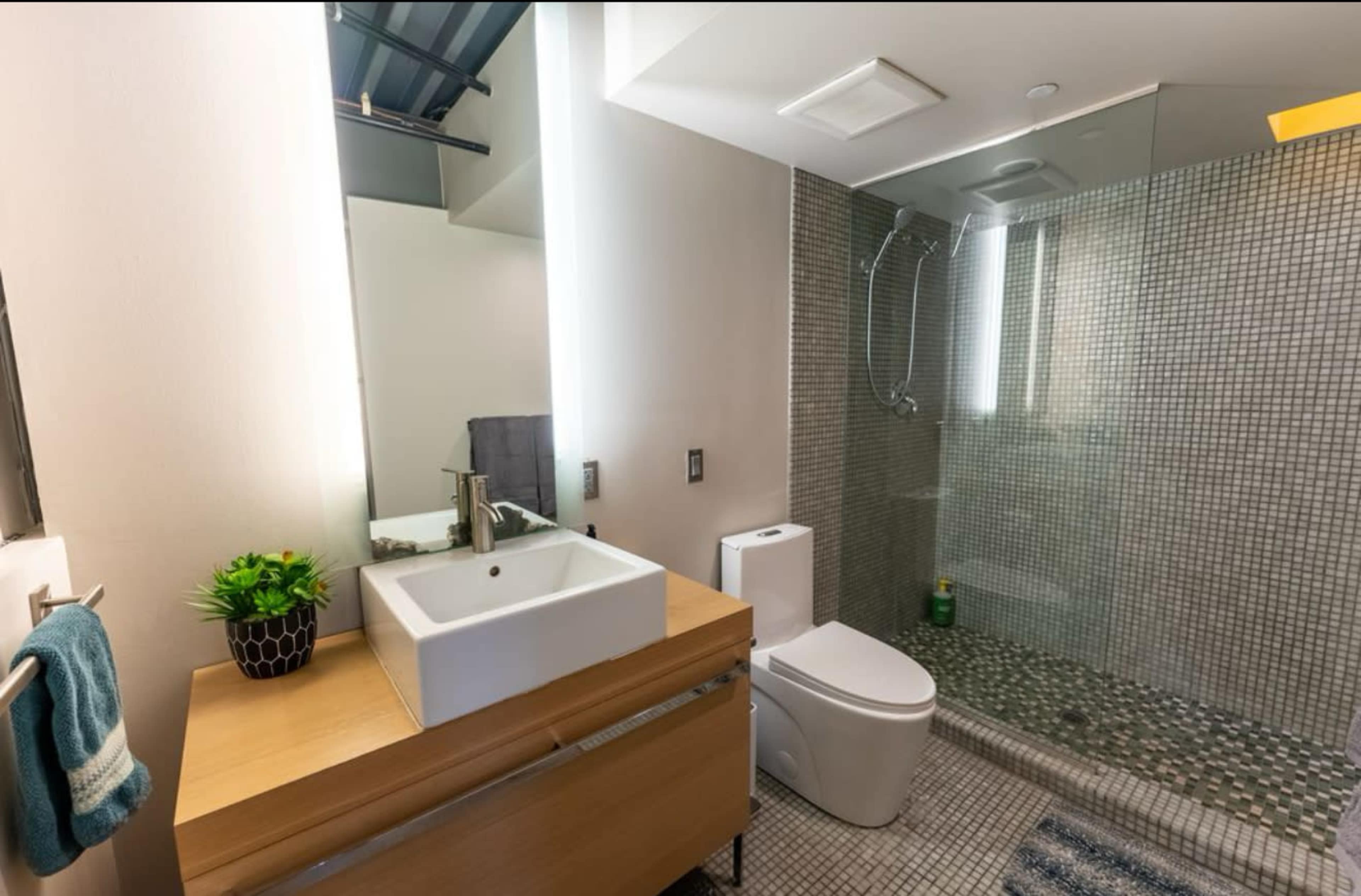 The image shows a modern bathroom featuring a glass shower, a wall-mounted sink with a wooden vanity, and a toilet, all accented by tile work and ambient lighting.