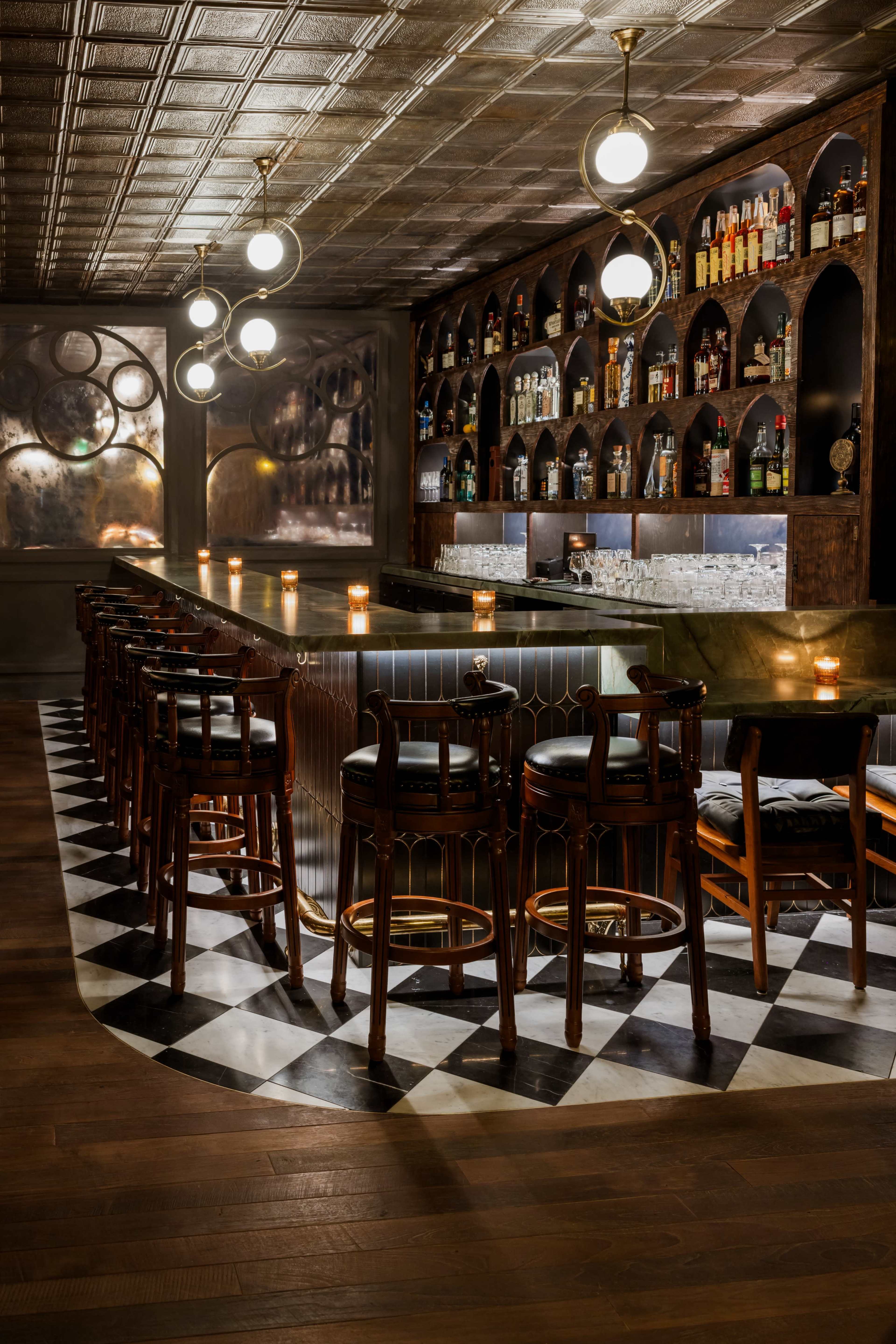 The image shows a dimly lit bar featuring a long, curved counter, high wooden stools, and shelves lined with various bottles against a backdrop of patterned walls.