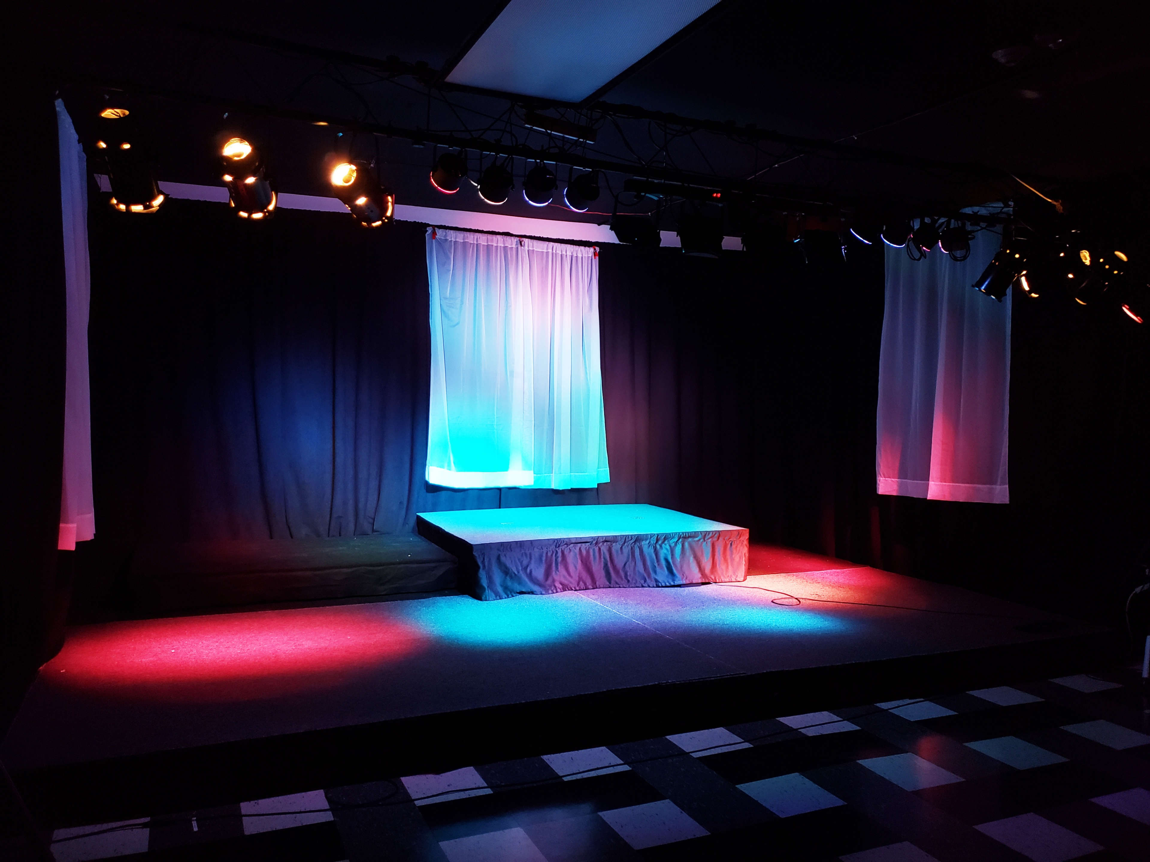 An empty stage with a raised platform, illuminated by colorful lights and flanked by white curtains.