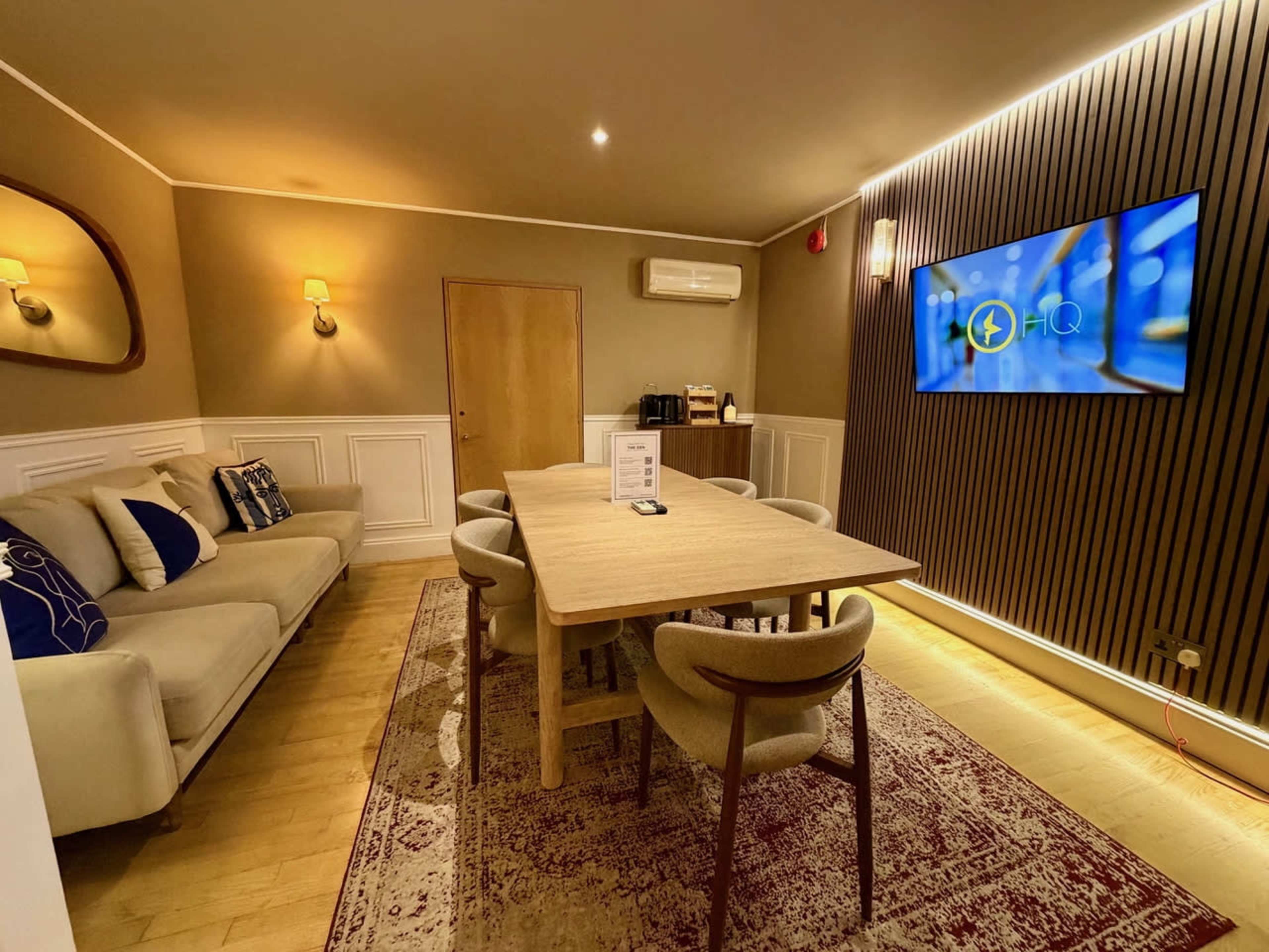 The image shows a well-furnished meeting room with a dining table surrounded by chairs, a sofa, and a large screen mounted on the wall.