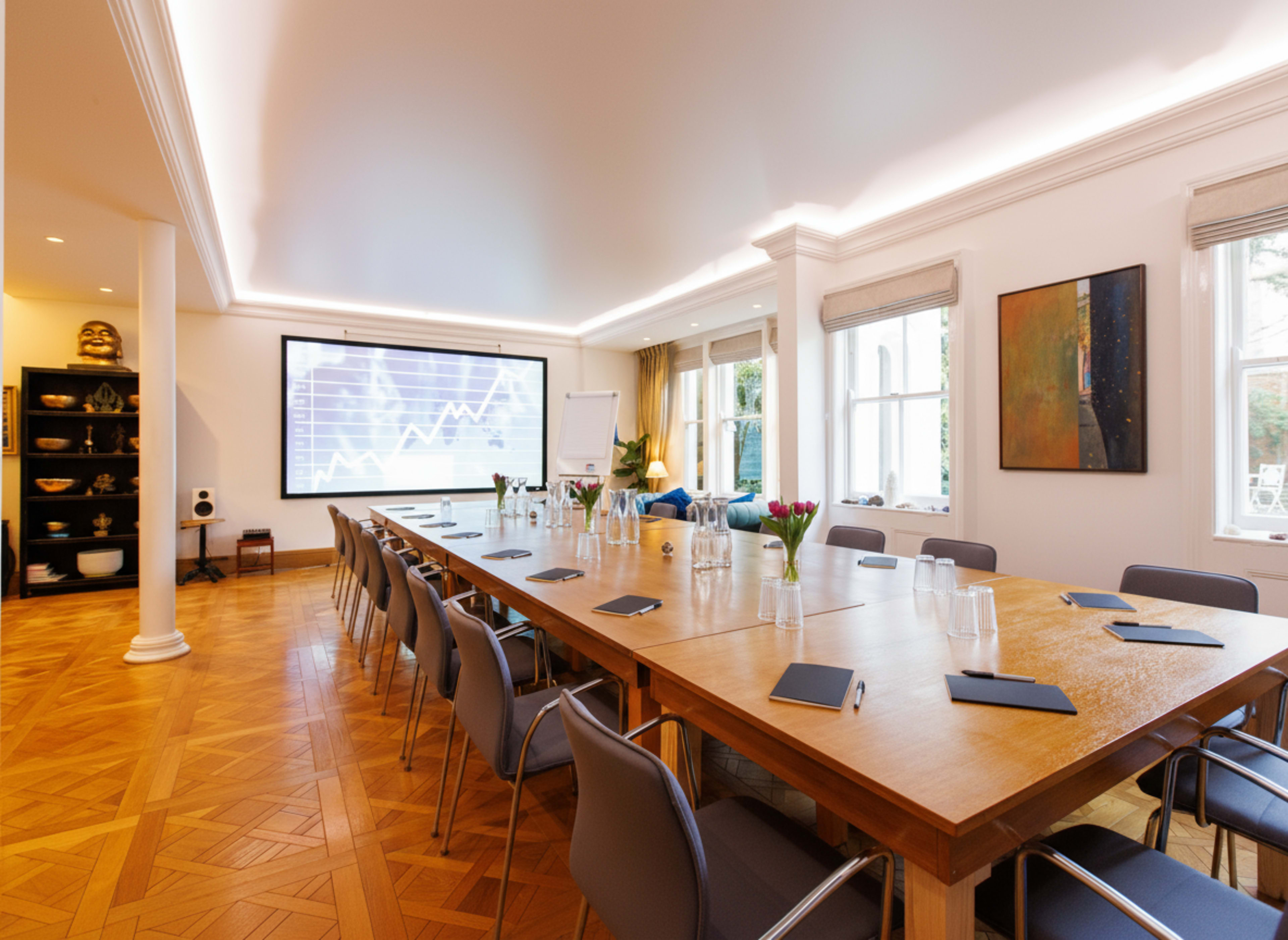 A modern conference room features a large wooden table surrounded by chairs, with a screen displaying a graph on one wall and large windows allowing natural light.