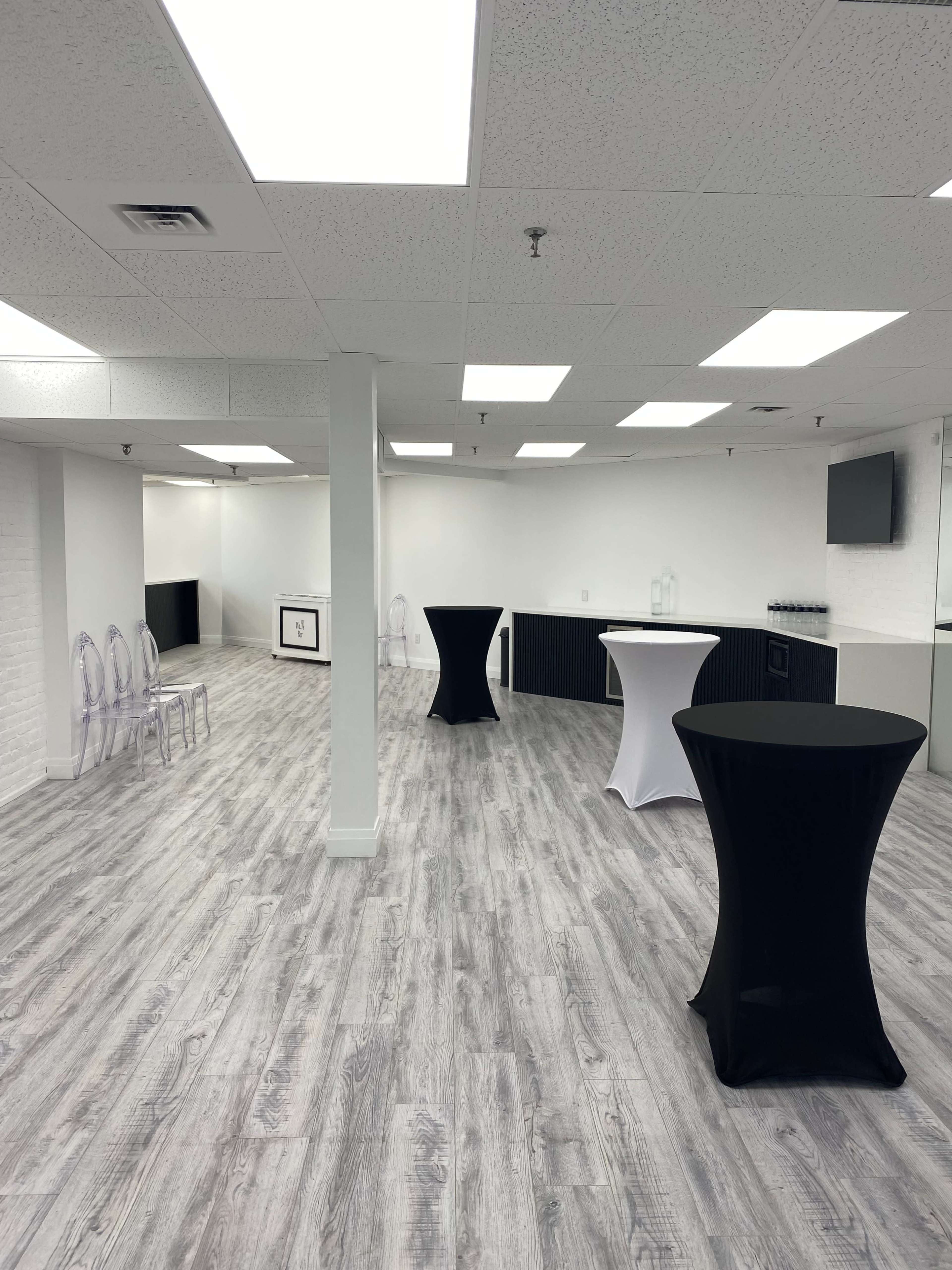 The image depicts a spacious, well-lit room with a light wood floor, featuring several high tables and a minimalist bar area along one wall.