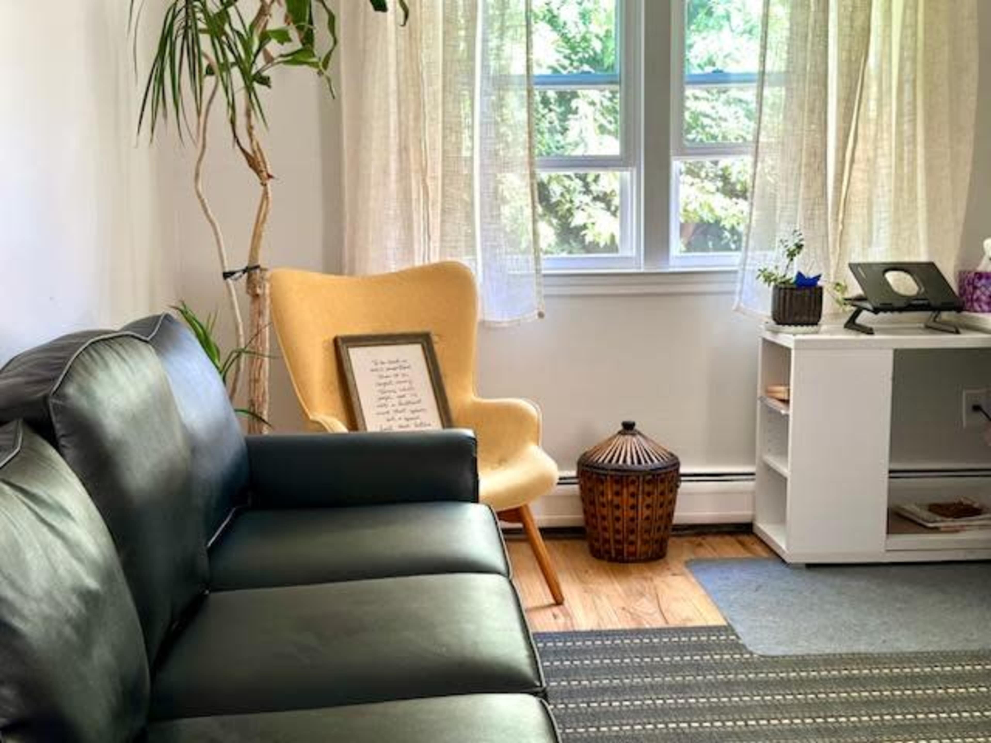 A living room features a black leather sofa, a yellow accent chair, a small wooden side table, and large windows with sheer curtains allowing natural light to enter.