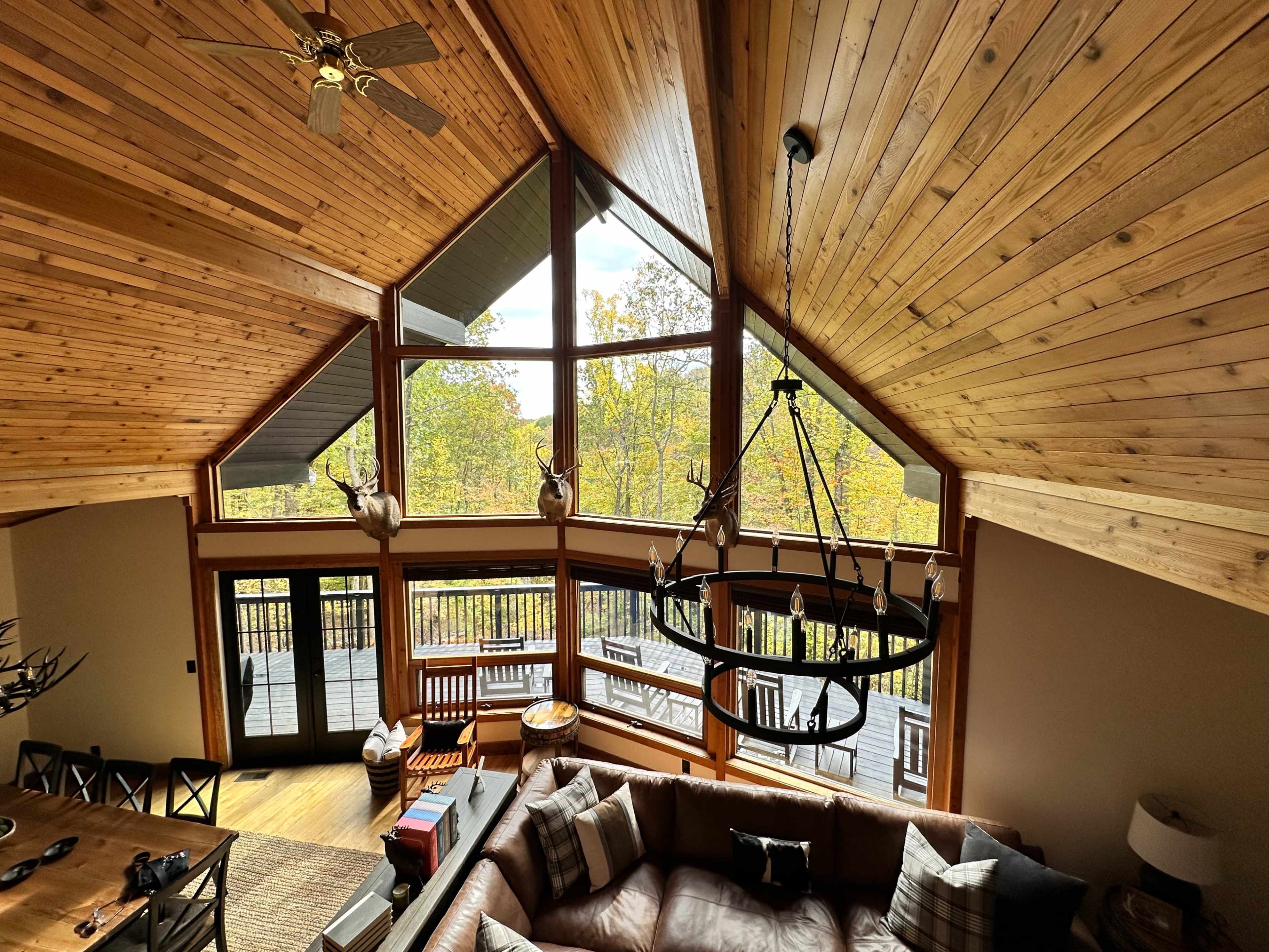 The image shows a spacious interior of a home's living area with high, wooden ceilings and large windows showcasing a view of trees outside.