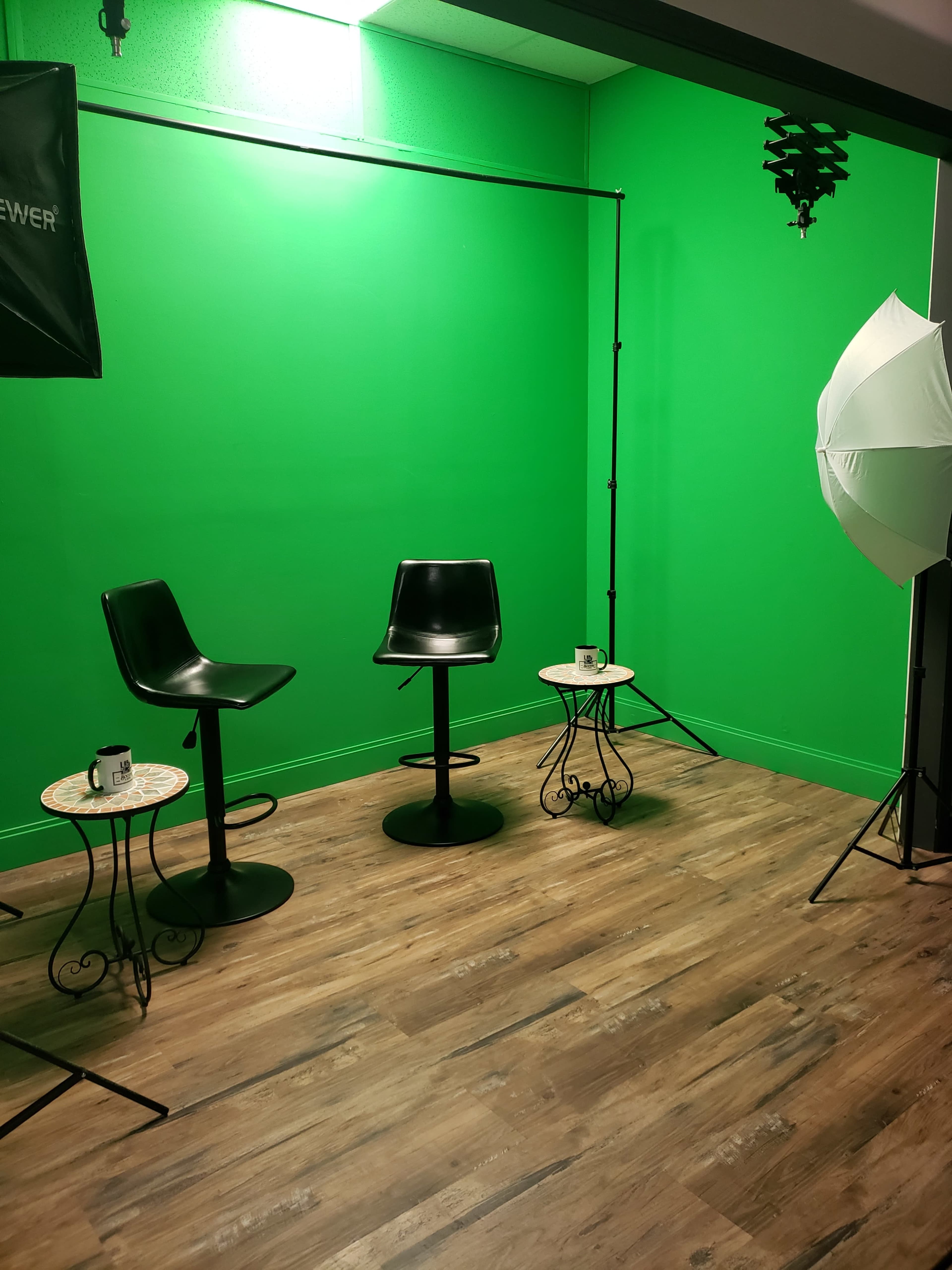 The image shows a small studio space with a bright green backdrop, two black stools, two small tables, and lighting equipment.