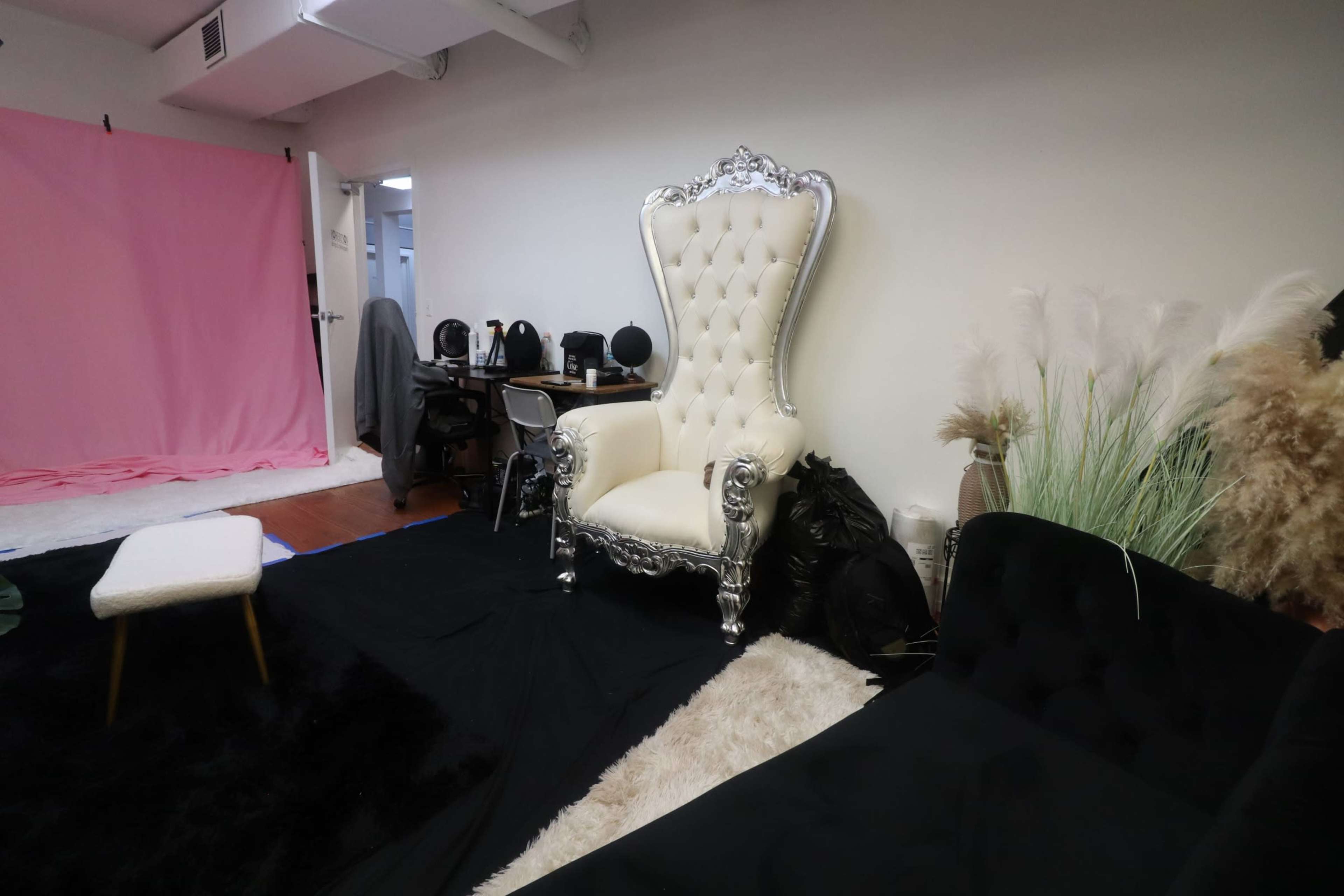 The image shows a spacious room with a white throne-like chair, a pink backdrop, various black furniture pieces, and decorative plants.