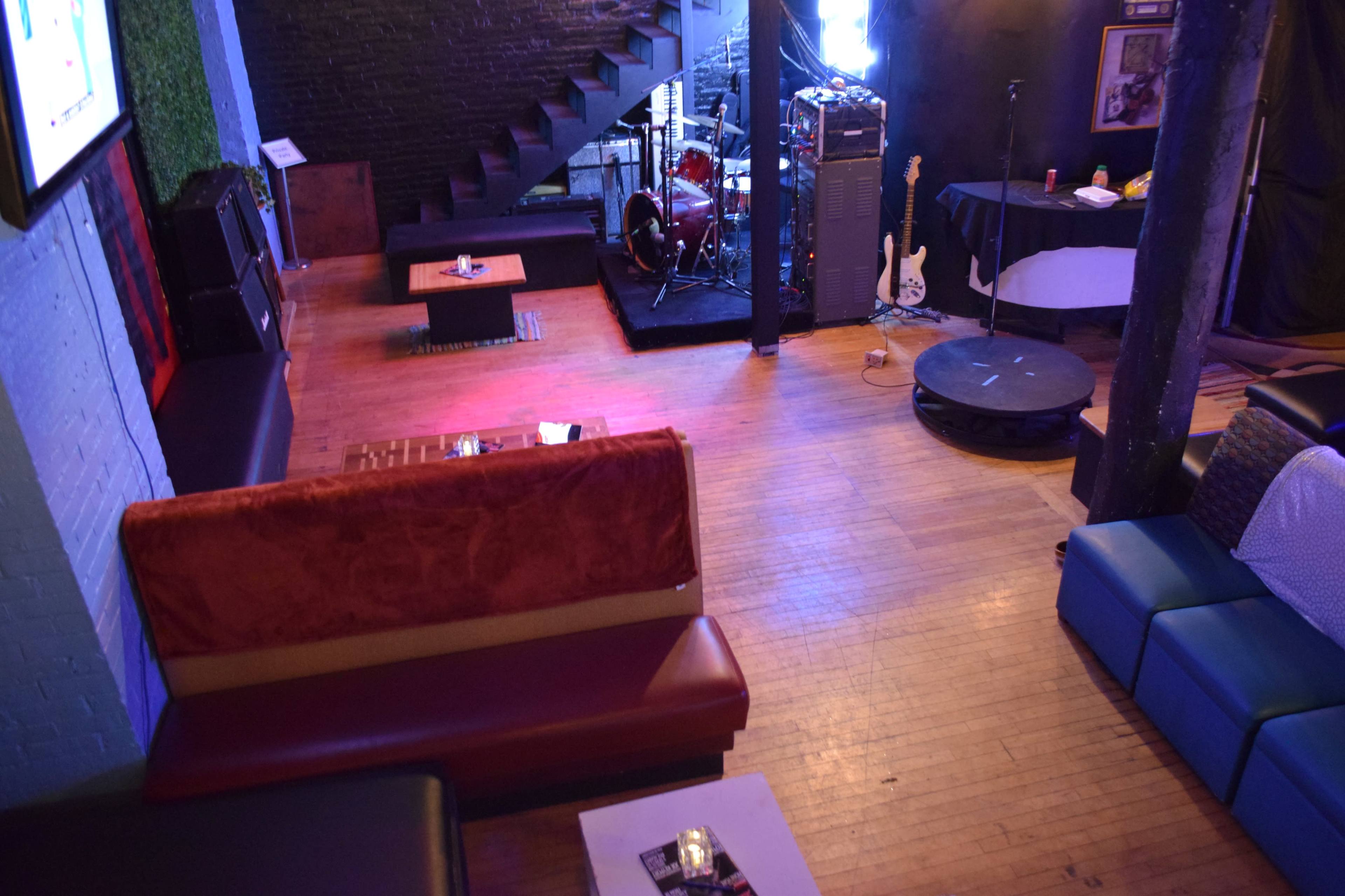 The image shows a music venue with a stage set up for a band, featuring a seating area with couches and a wooden floor.