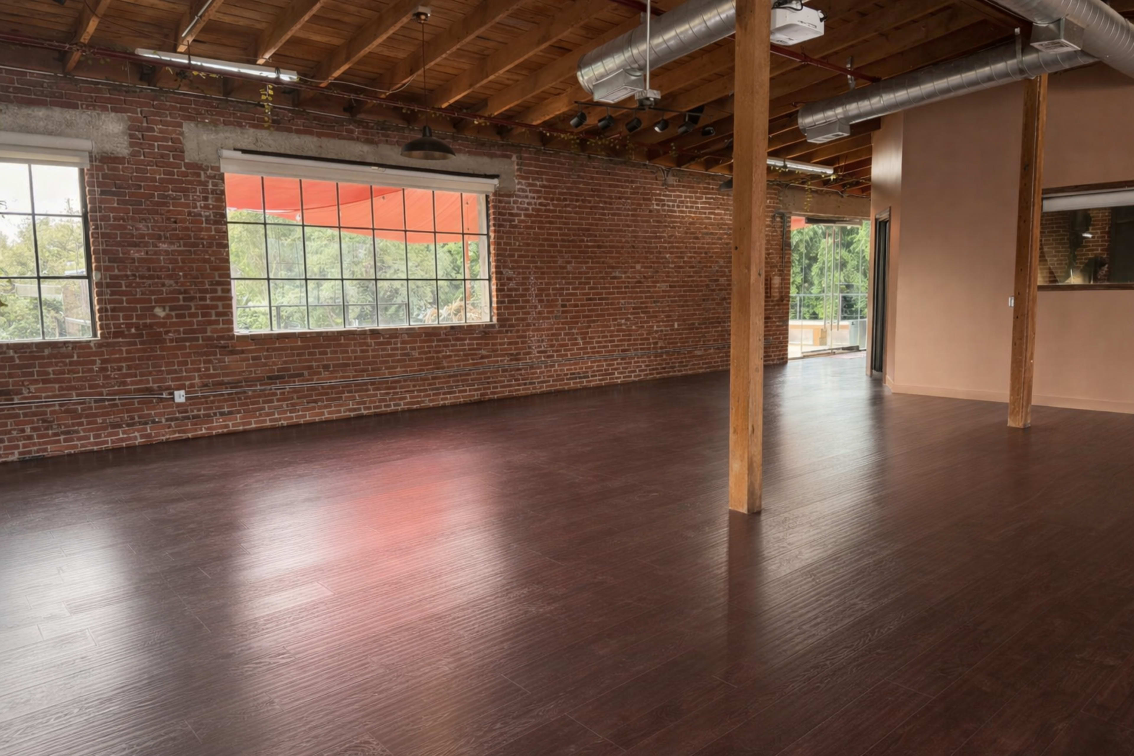 The image shows a spacious, empty room with exposed brick walls, large windows, wooden beams, and a polished dark floor.