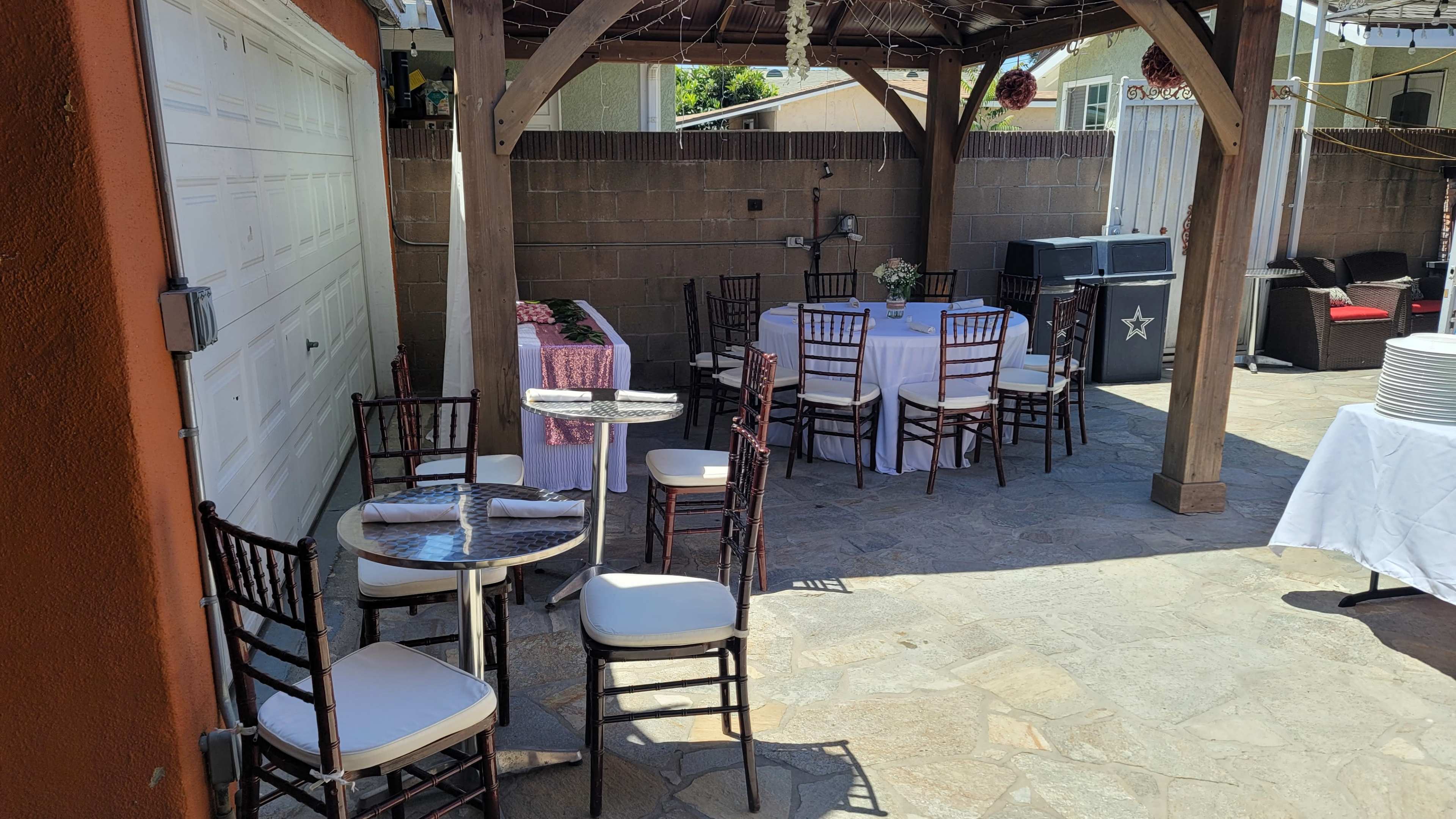 A patio area is set up with several tables and chairs under a wooden gazebo, featuring a stone floor and various decorations.