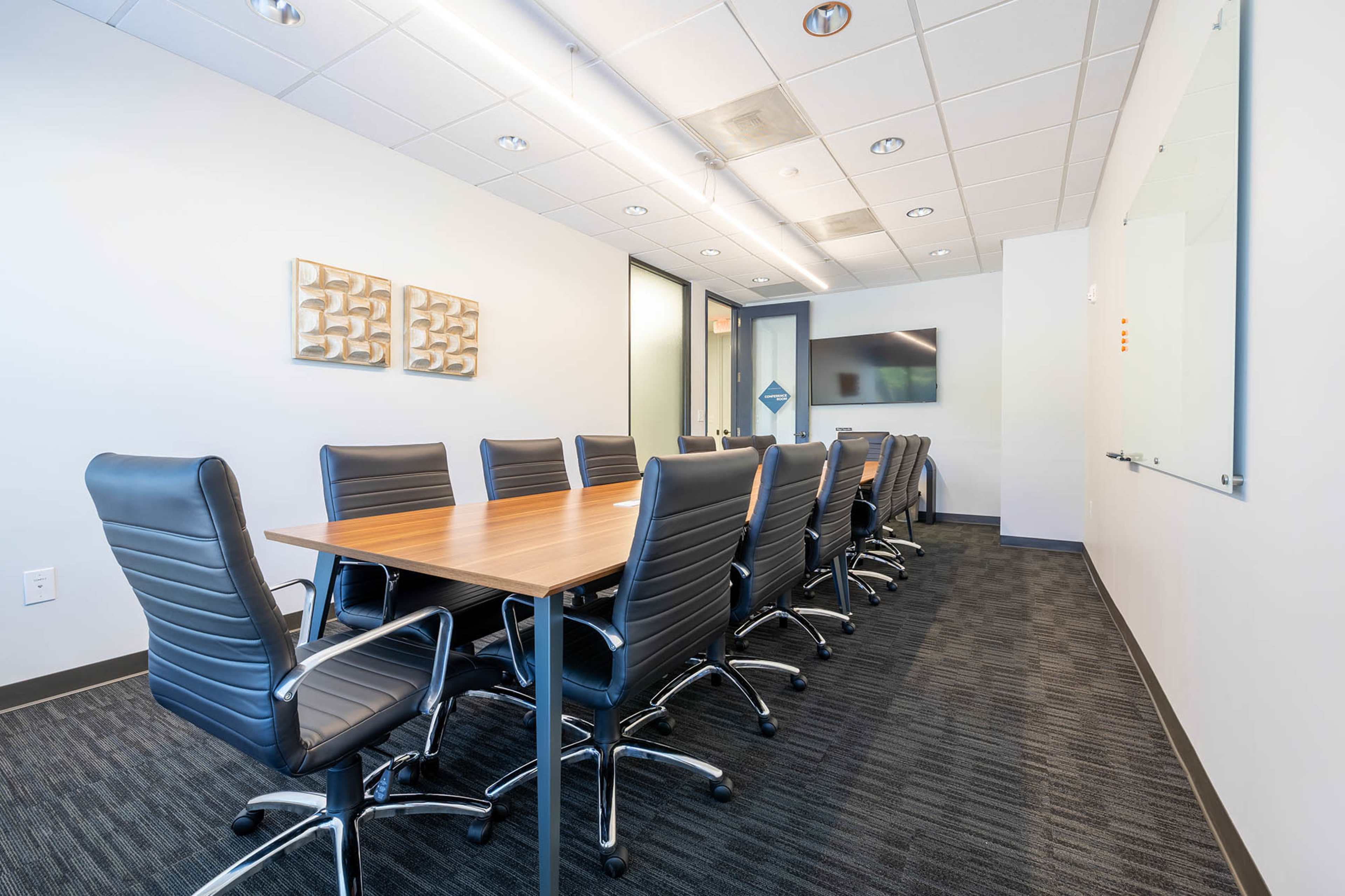 A modern conference room features a long wooden table surrounded by black leather chairs, with wall art and a large screen visible.