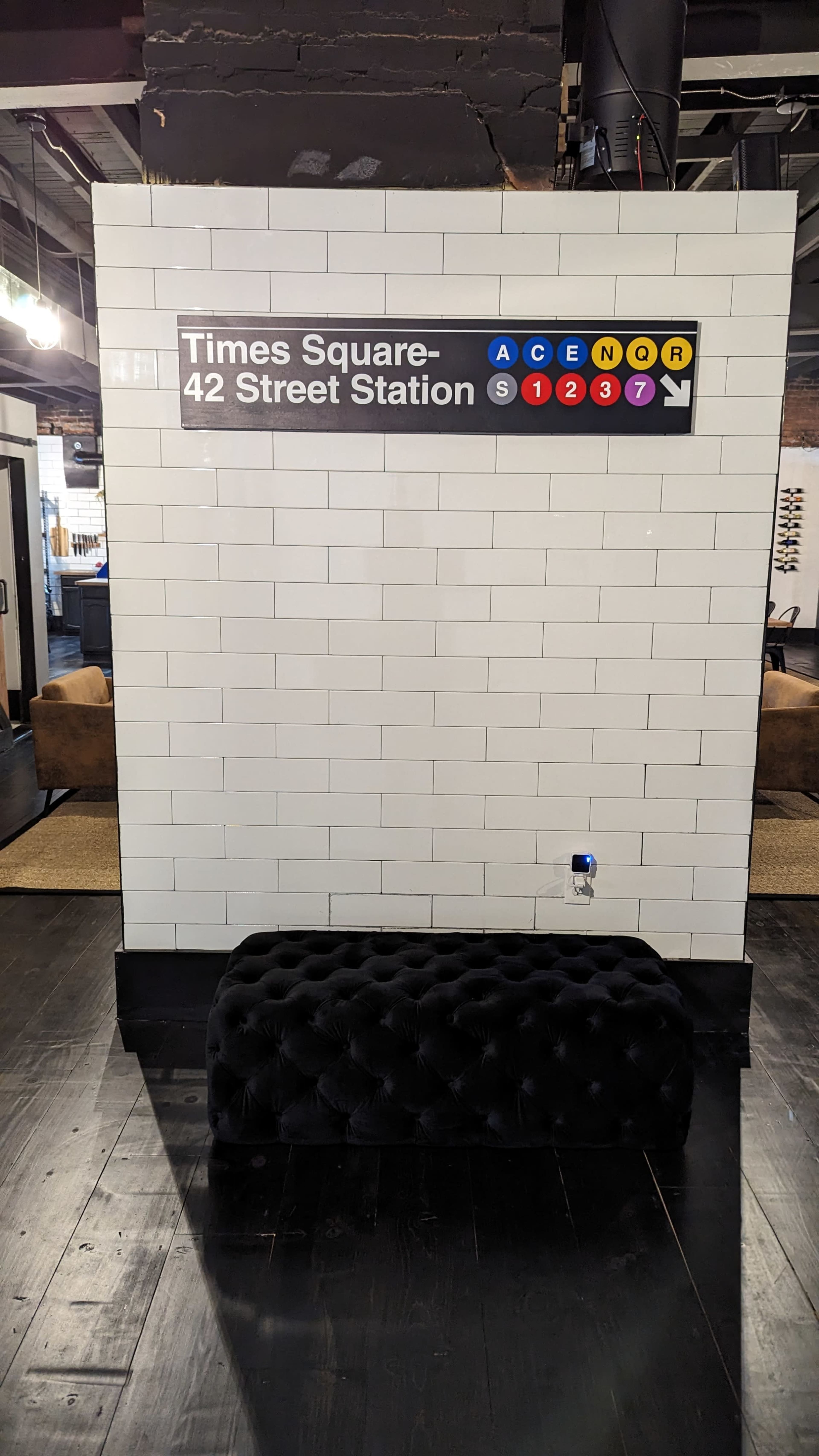 A tiled wall displaying a sign for "Times Square-42 Street Station" alongside various subway line symbols, with a black upholstered bench in front of it.