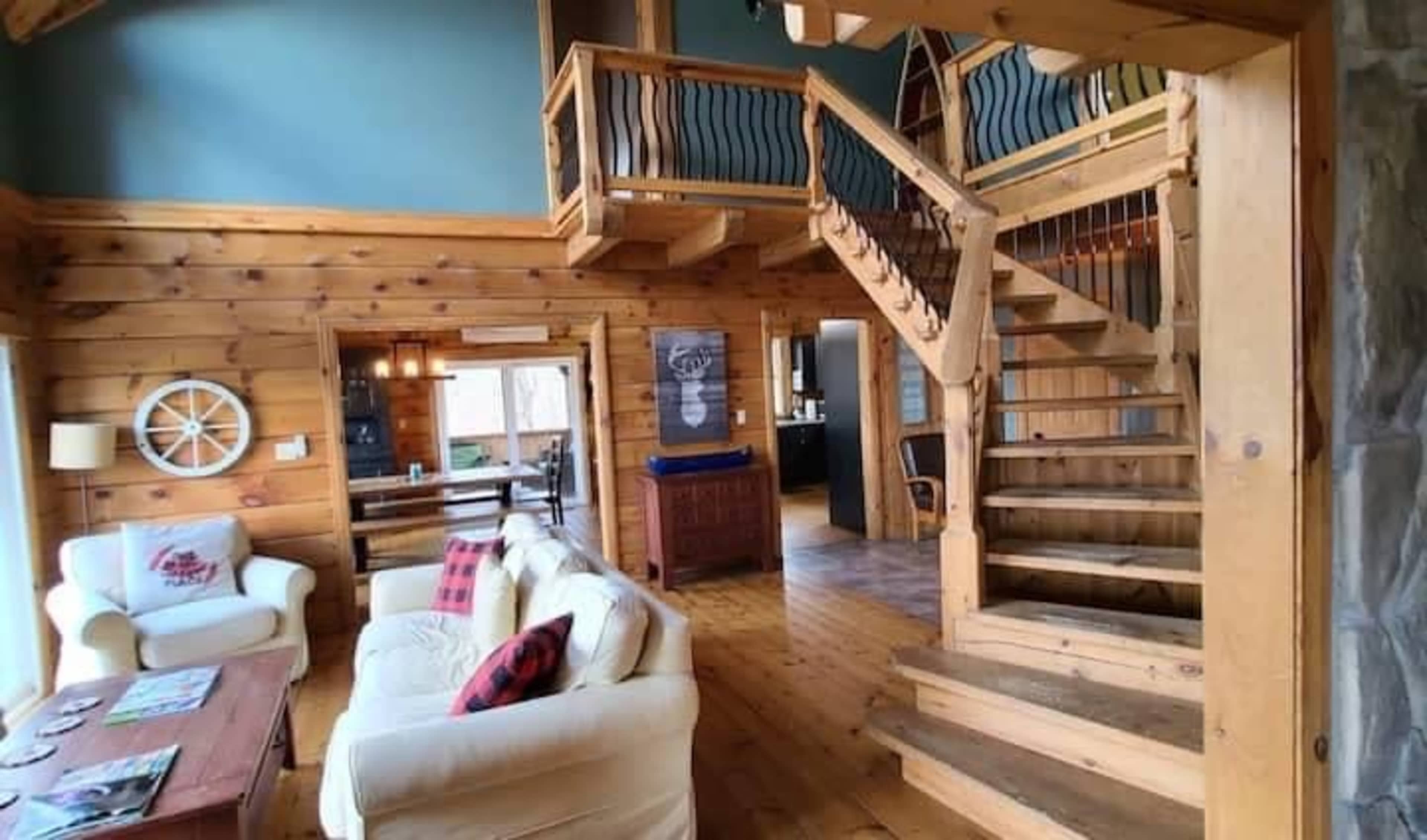 A wooden interior of a multi-level cabin, featuring a staircase, cozy seating area, and large windows.