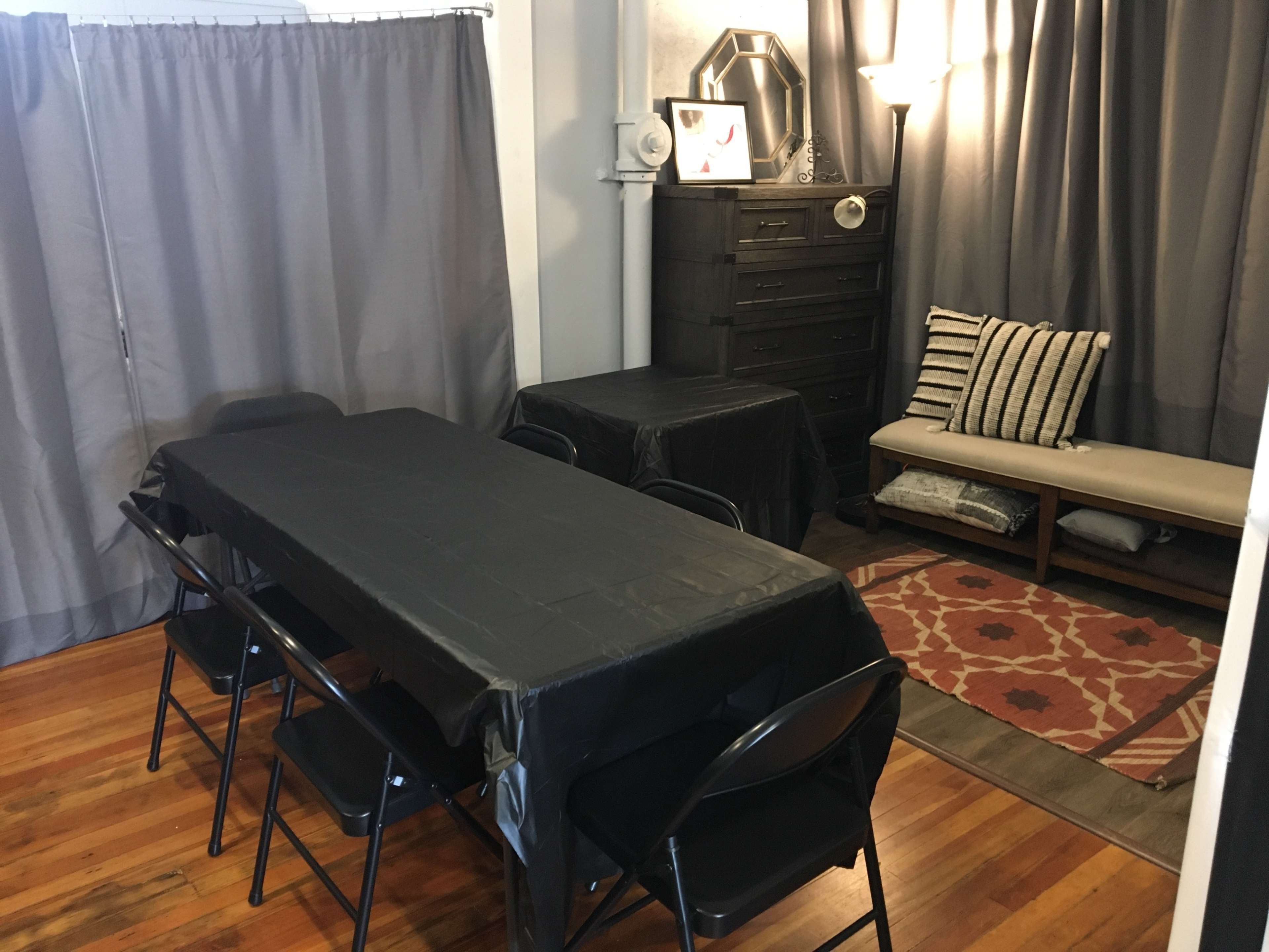 A dining area with a long black table surrounded by folding chairs, a dresser, and a bench with pillows, all set against gray curtains and wooden flooring.