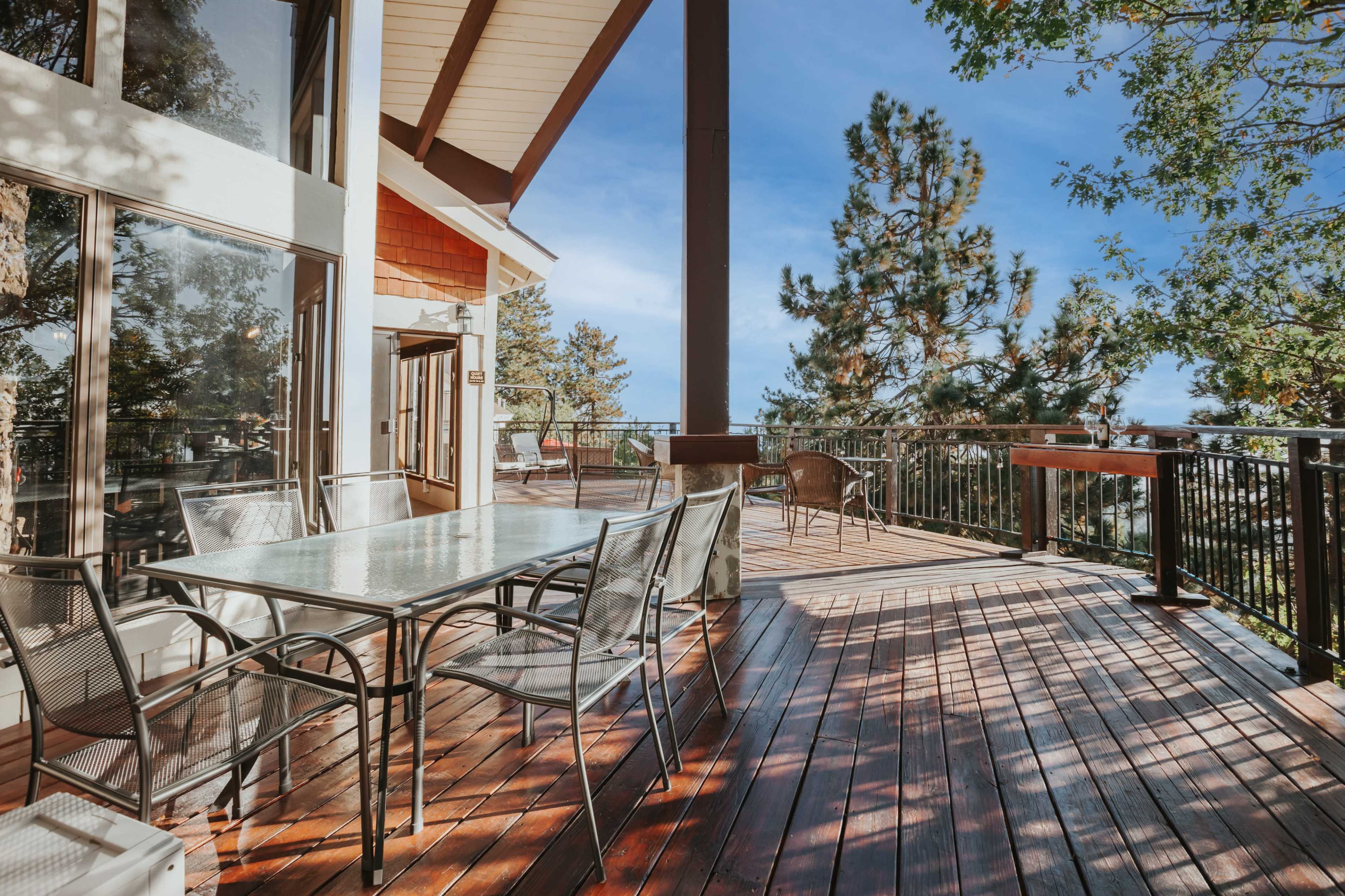 A spacious outdoor deck features a glass-top table surrounded by metal chairs, overlooking a treed landscape under a clear sky.