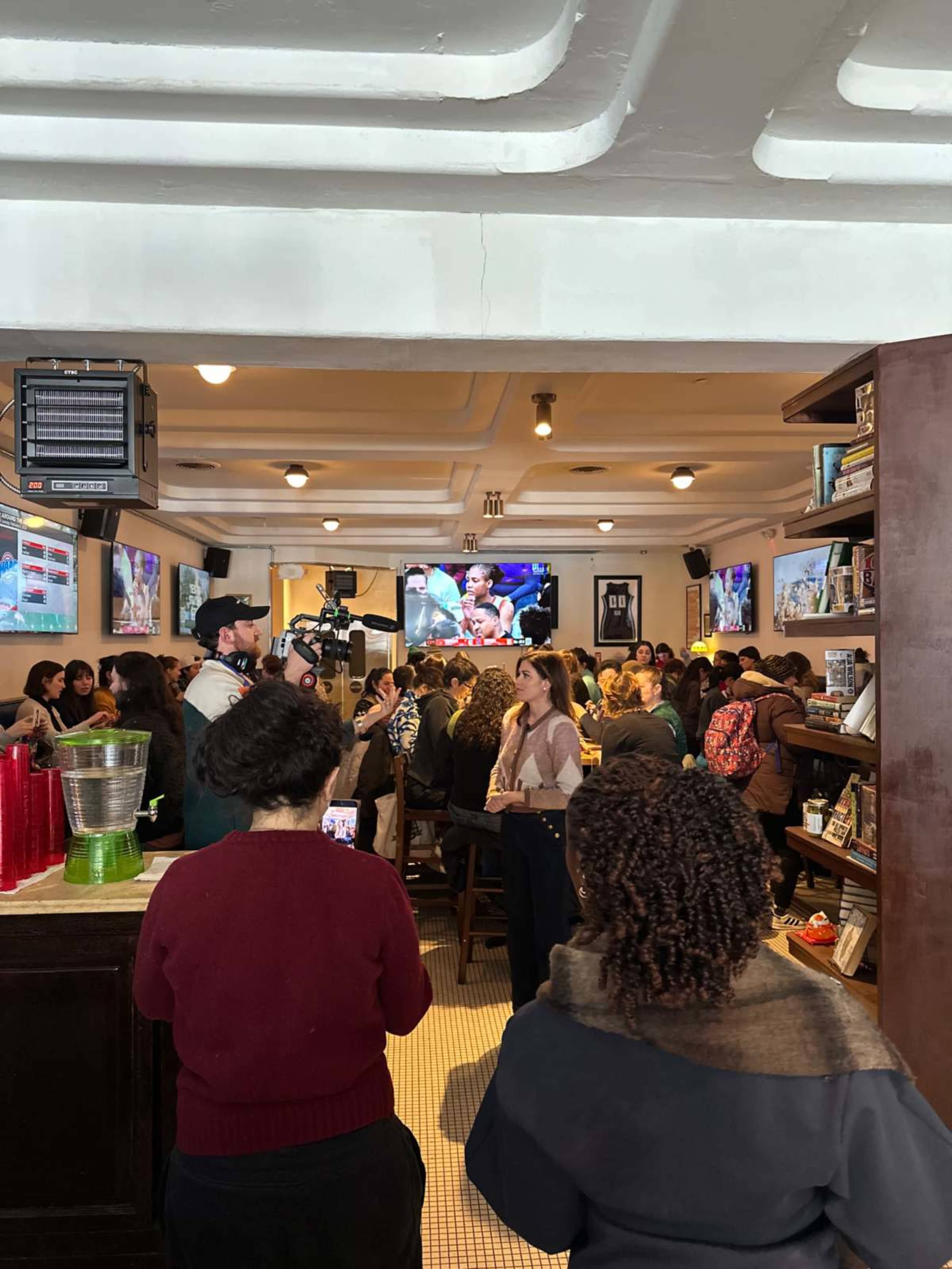 A crowded bar is filled with people watching a televised event while others engage in conversation.