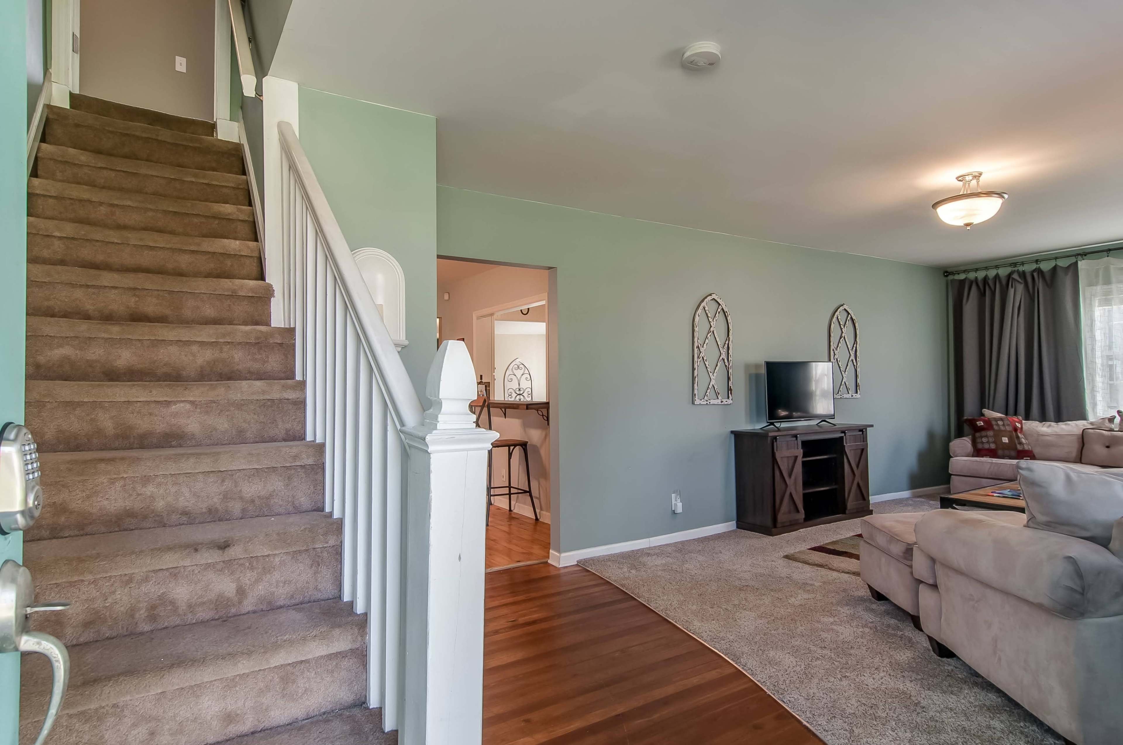 A light-filled living area with a staircase leading to the upper floor, featuring a TV stand, couch, and a view into an adjoining room.