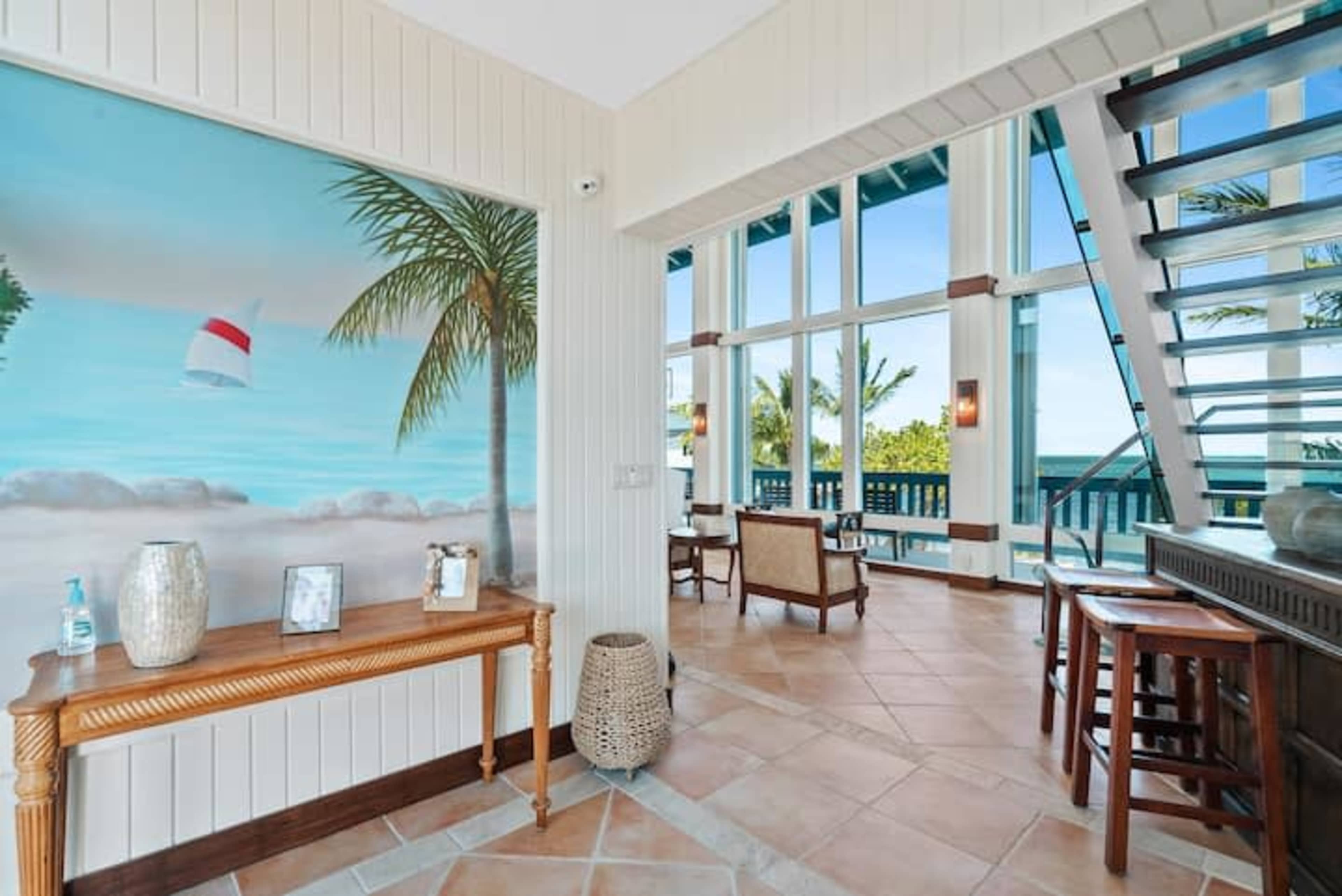 The image shows a well-lit interior featuring a coastal-themed mural, wooden furniture, and large windows offering a view of the ocean.