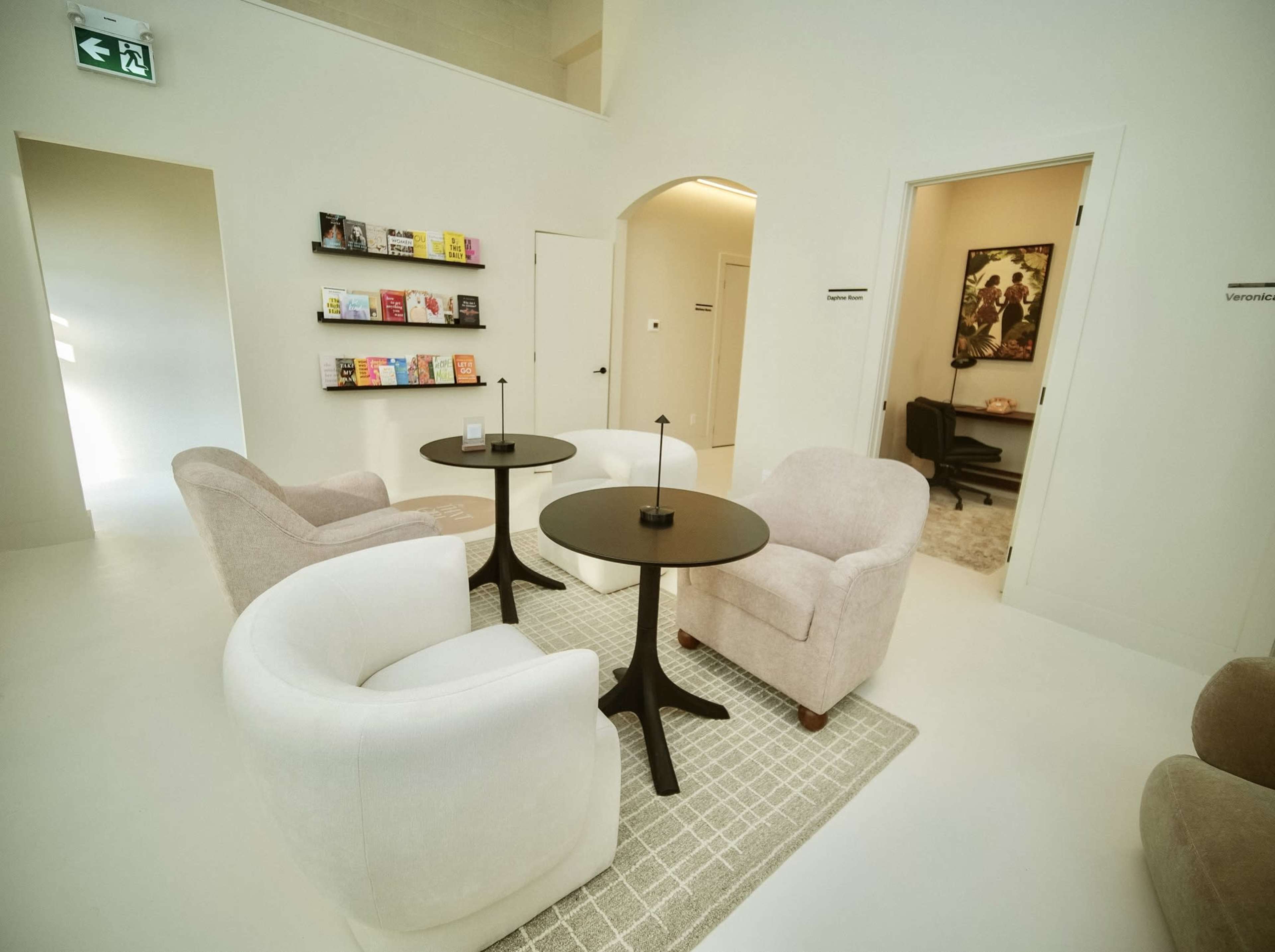 The image shows a minimalistic lounge area featuring two round tables surrounded by four upholstered chairs, with a bookshelf displaying magazines in the background.
