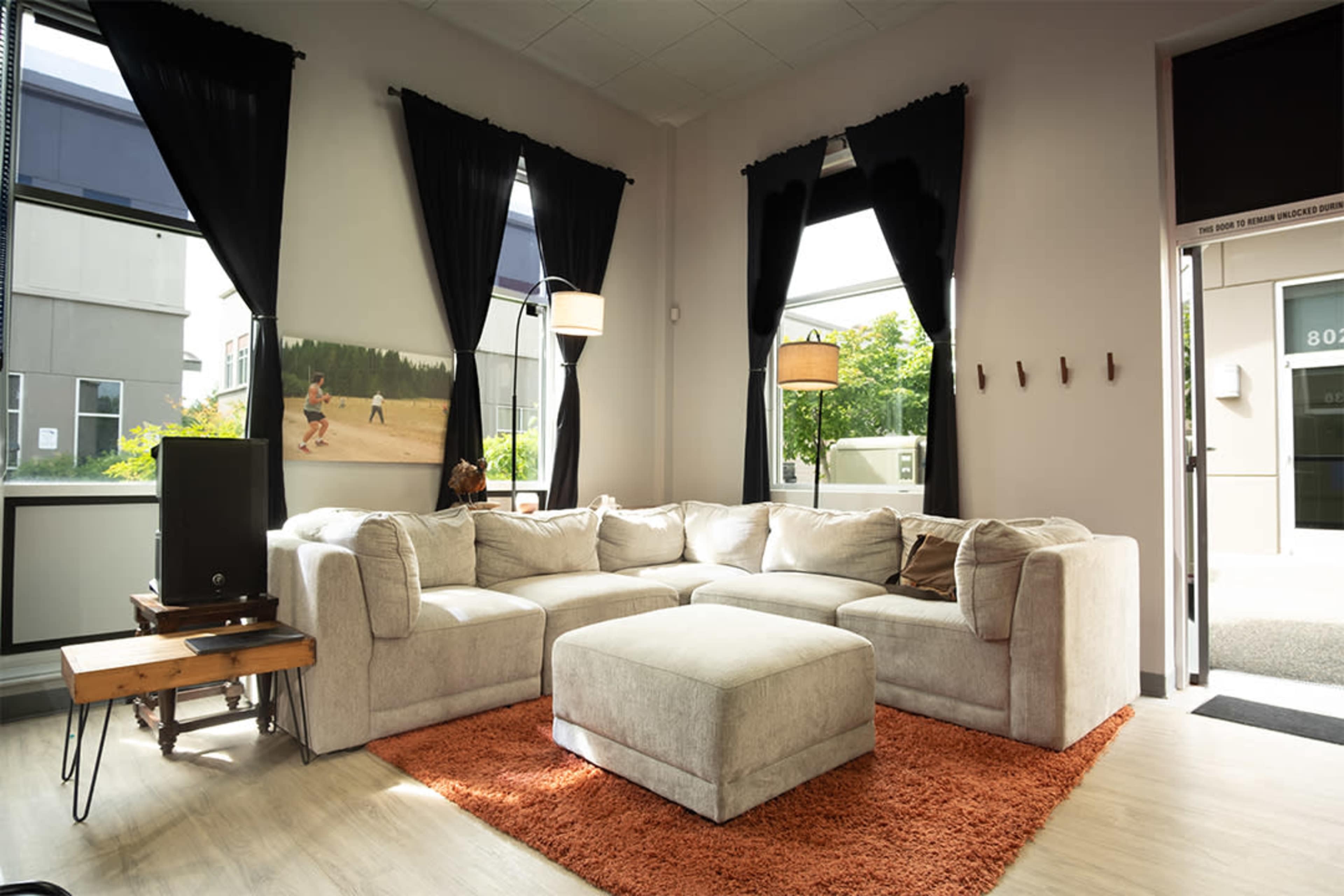 The image shows a cozy living room with a large, light-colored sectional sofa, an orange rug, and windows adorned with black curtains, allowing natural light to fill the space.