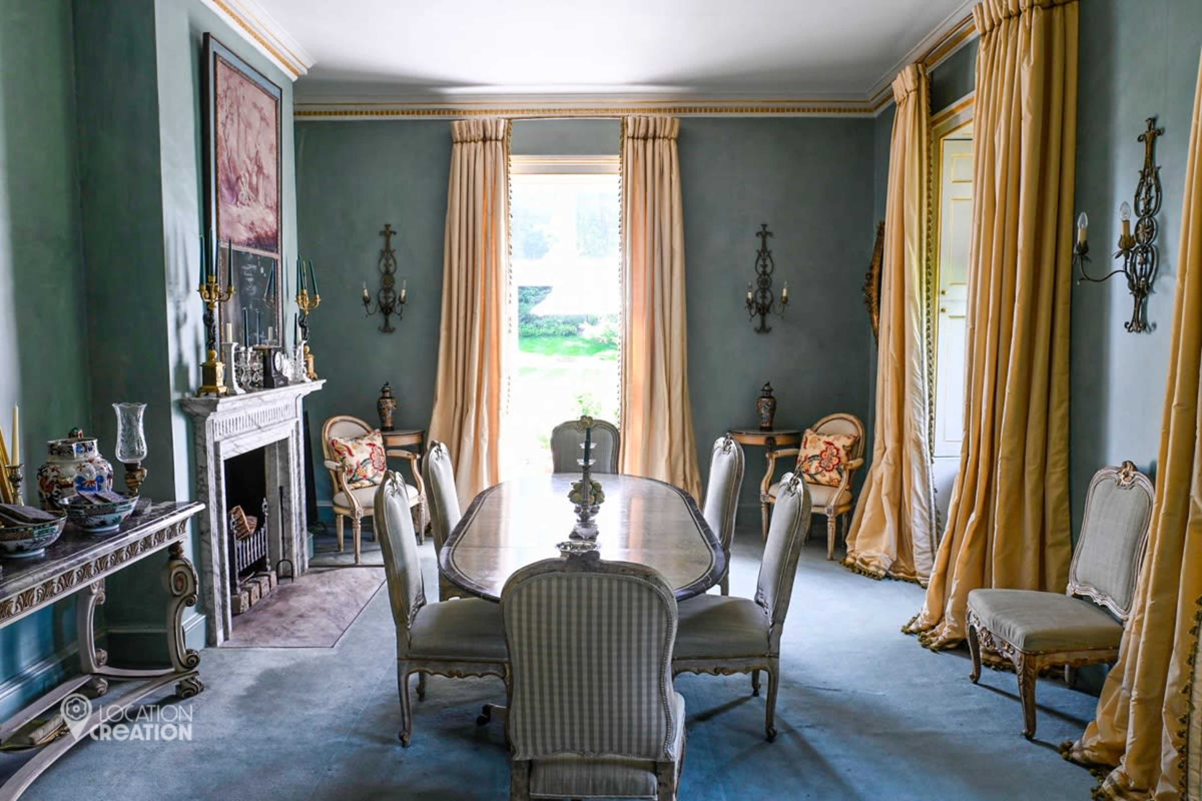 The image shows a dining room with a long table surrounded by chairs, adorned with elegant curtains and a fireplace.