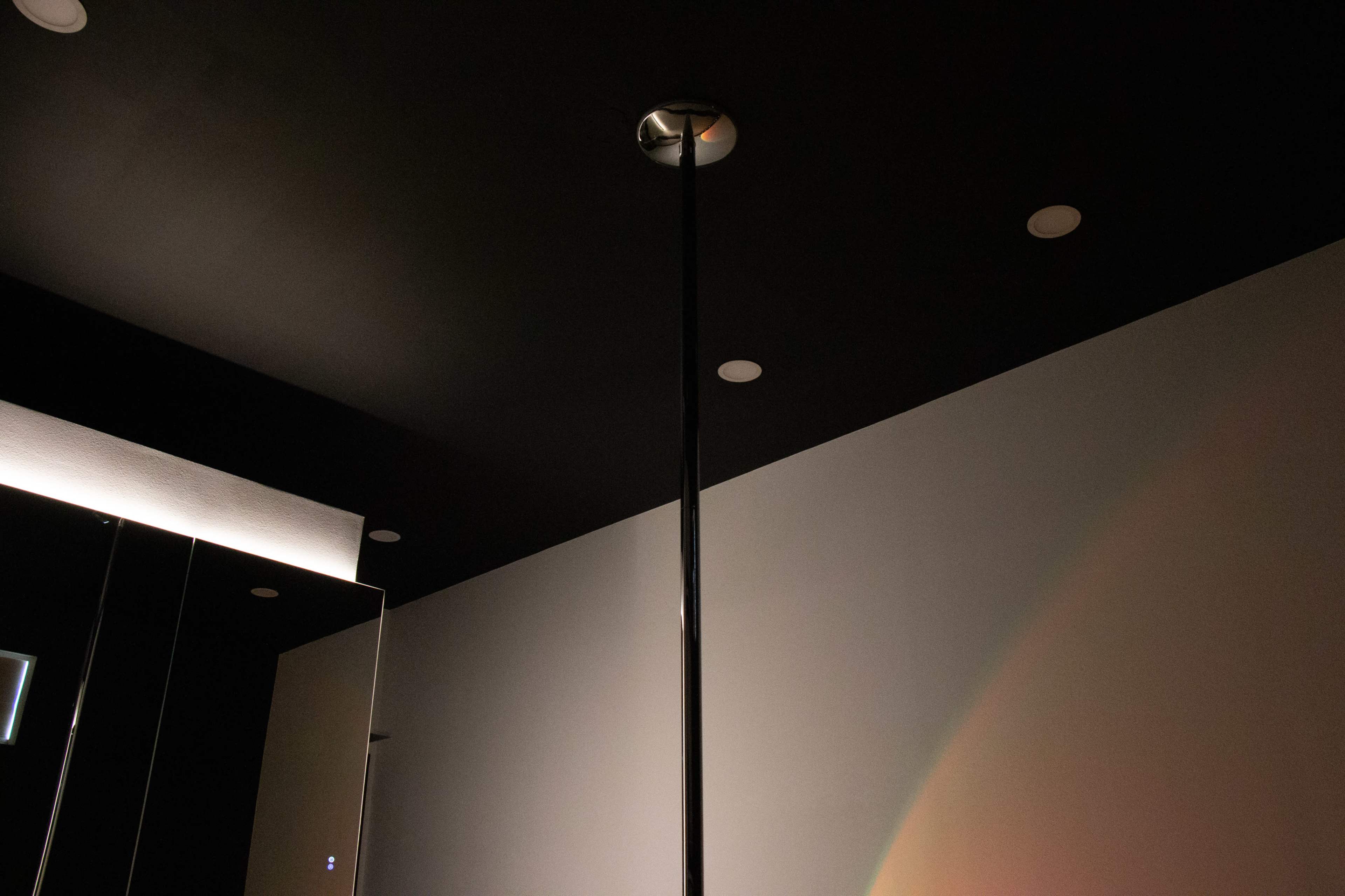 The image shows a dimly lit room with a black ceiling, featuring a vertical pole in the center and a mirror on one wall.