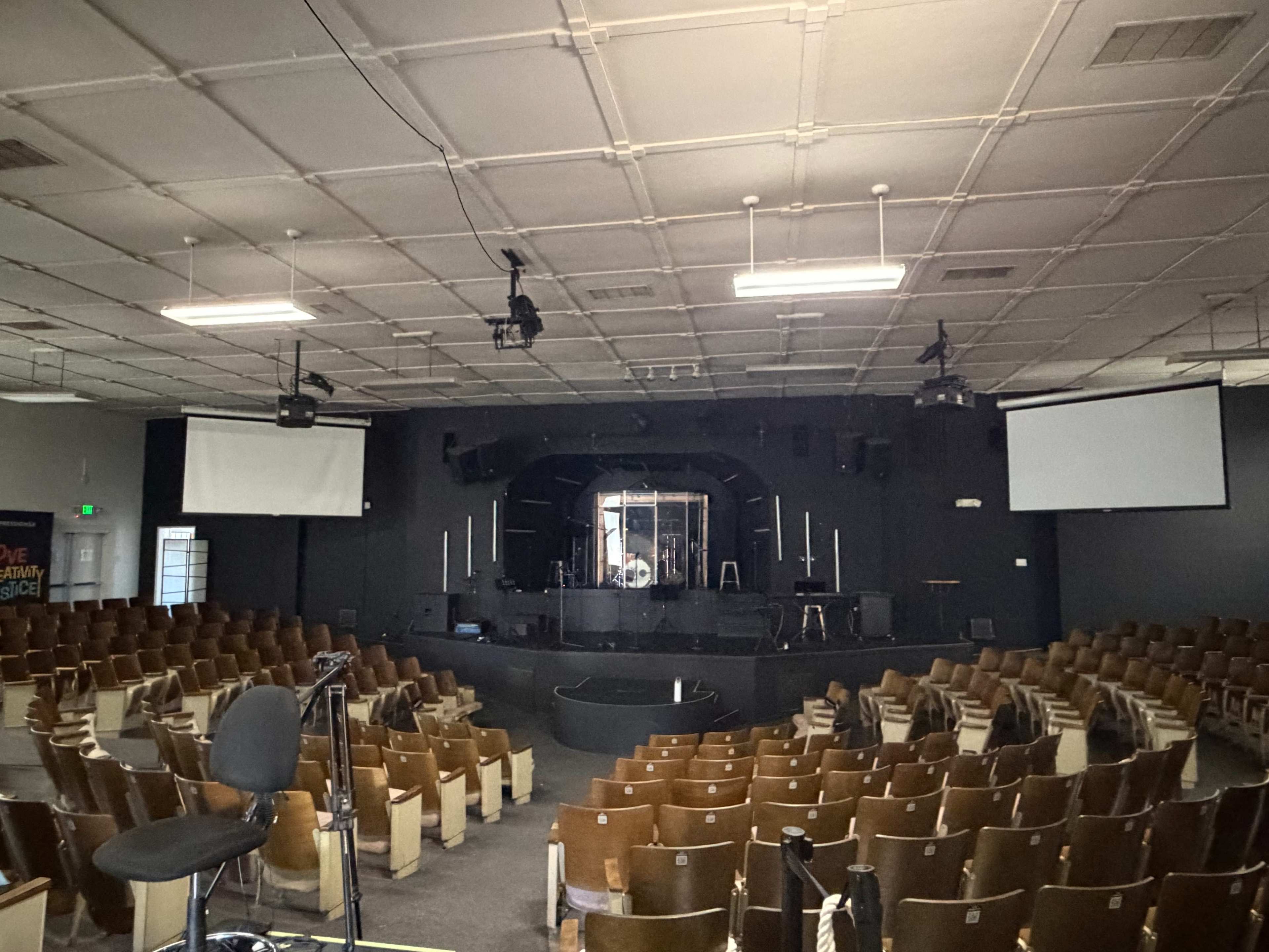 The image shows a large, nearly empty auditorium with tiered seating facing a stage that features various lighting and sound equipment.