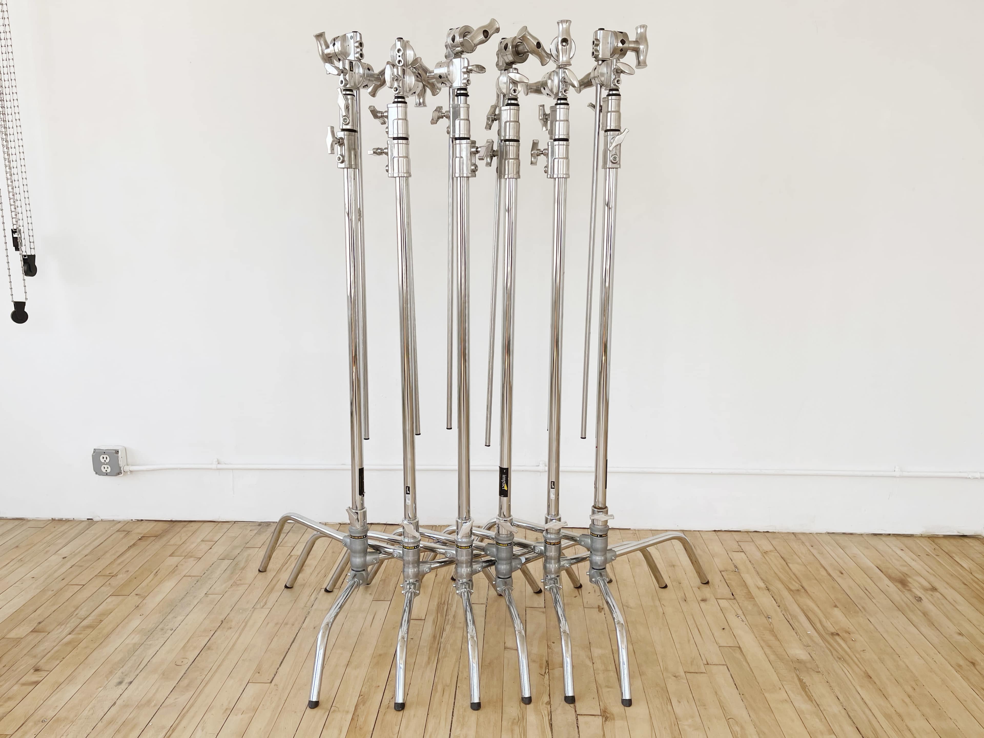 A group of six chrome light stands arranged in a row on a wooden floor in a plain white-walled space.