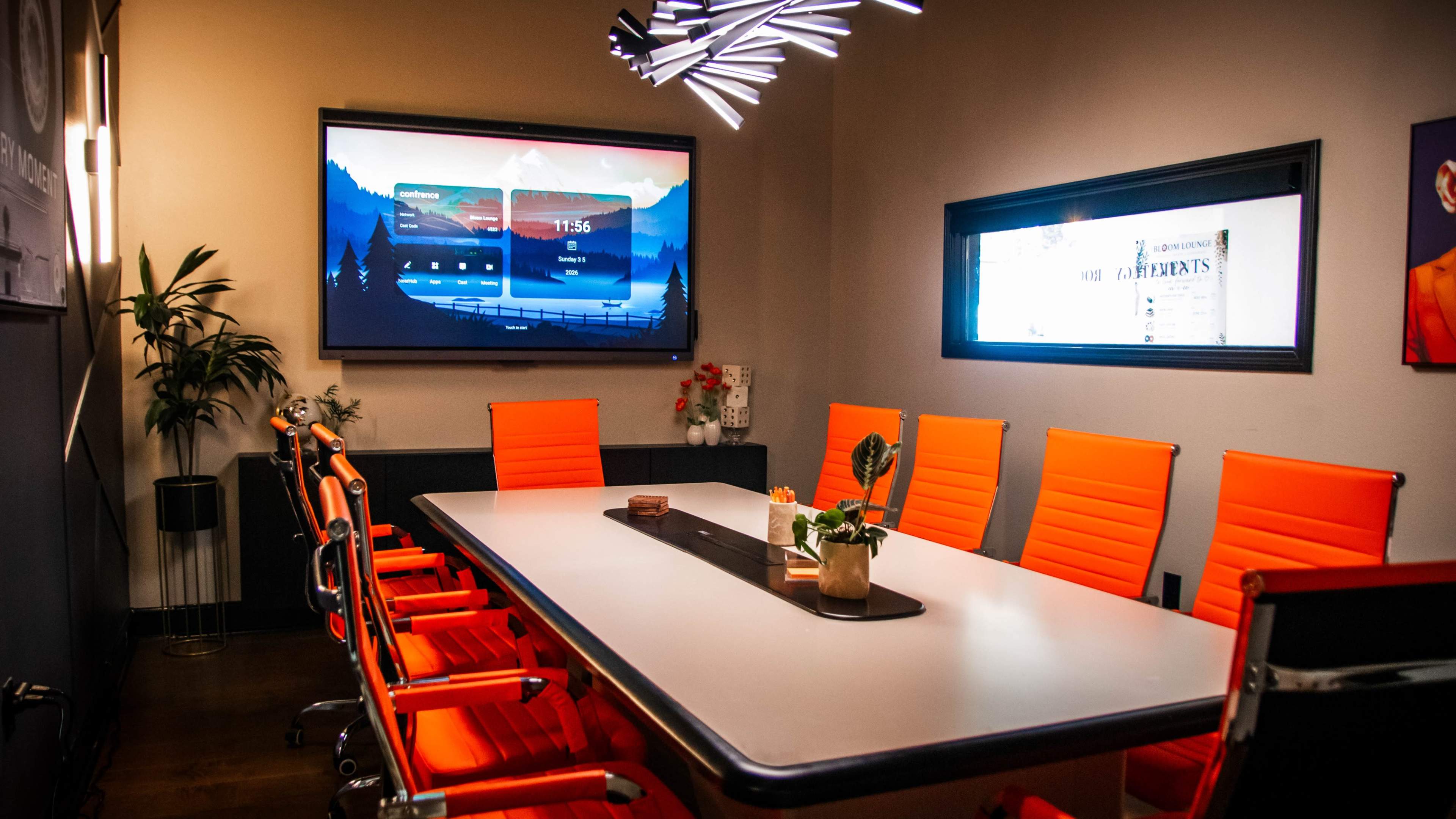 A modern conference room features a long white table surrounded by bright orange chairs and a large screen displaying a digital interface on the wall.