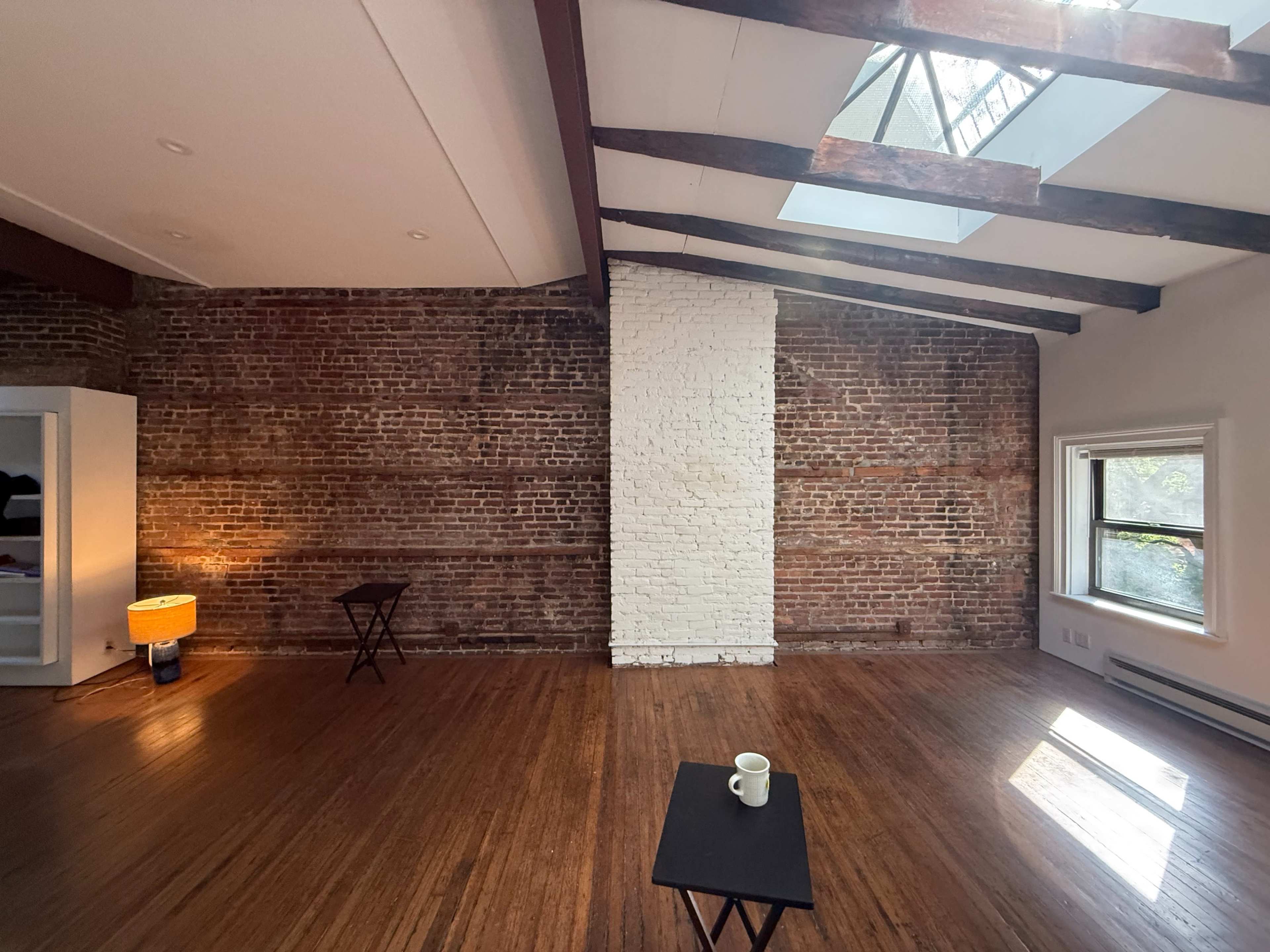 The image shows a spacious room with exposed brick walls, wooden beams, and a skylight, featuring minimal furniture.