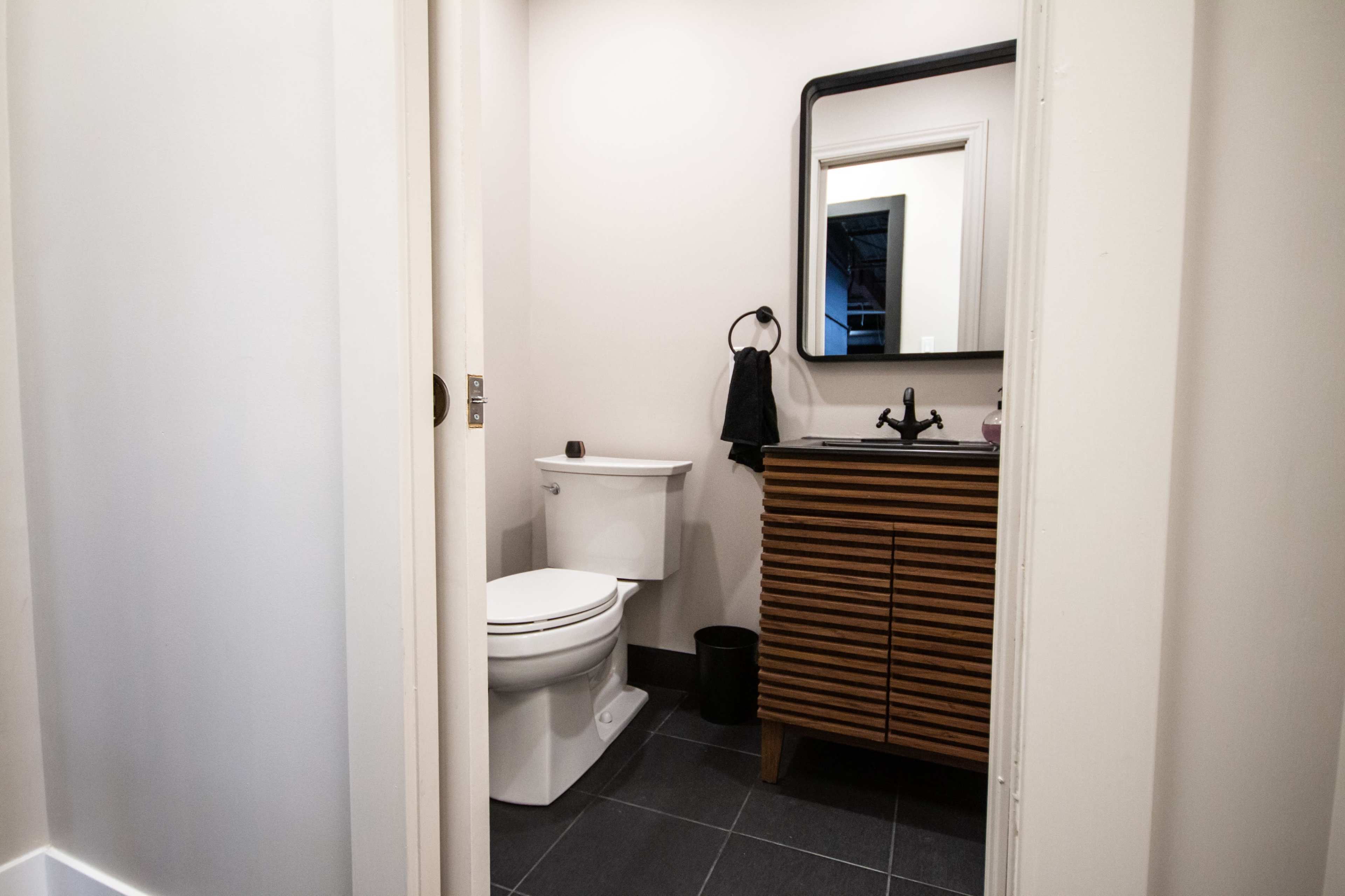A small bathroom features a toilet, a dark wooden vanity with a sink, and a mirror above it, all illuminated by natural light.