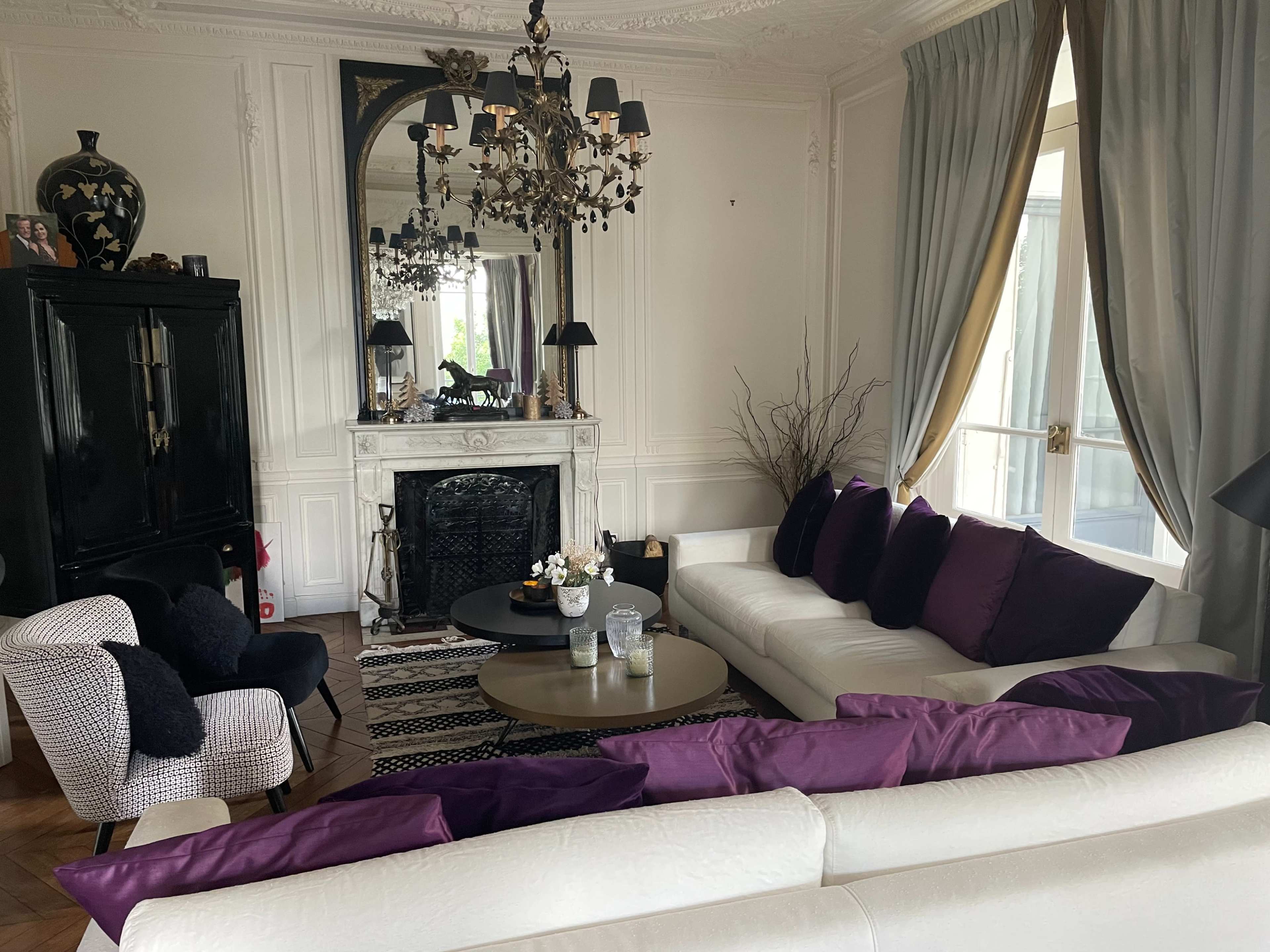 A elegantly styled living room featuring a white sofa with purple cushions, a marble fireplace, a large mirror, and a black side table.