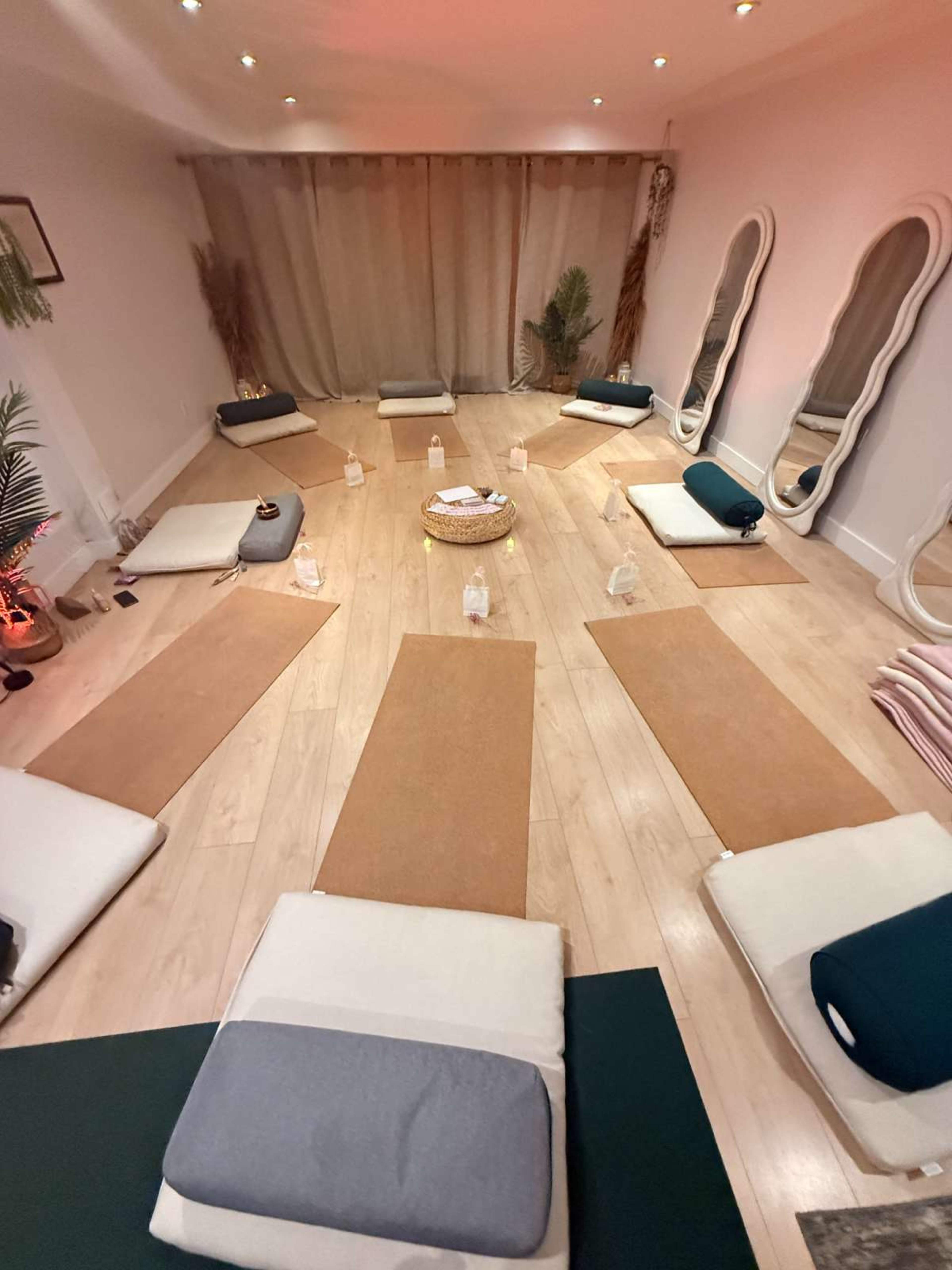 The image shows a yoga studio arranged with mats and cushions in circular formation, surrounded by soft lighting and mirrors.