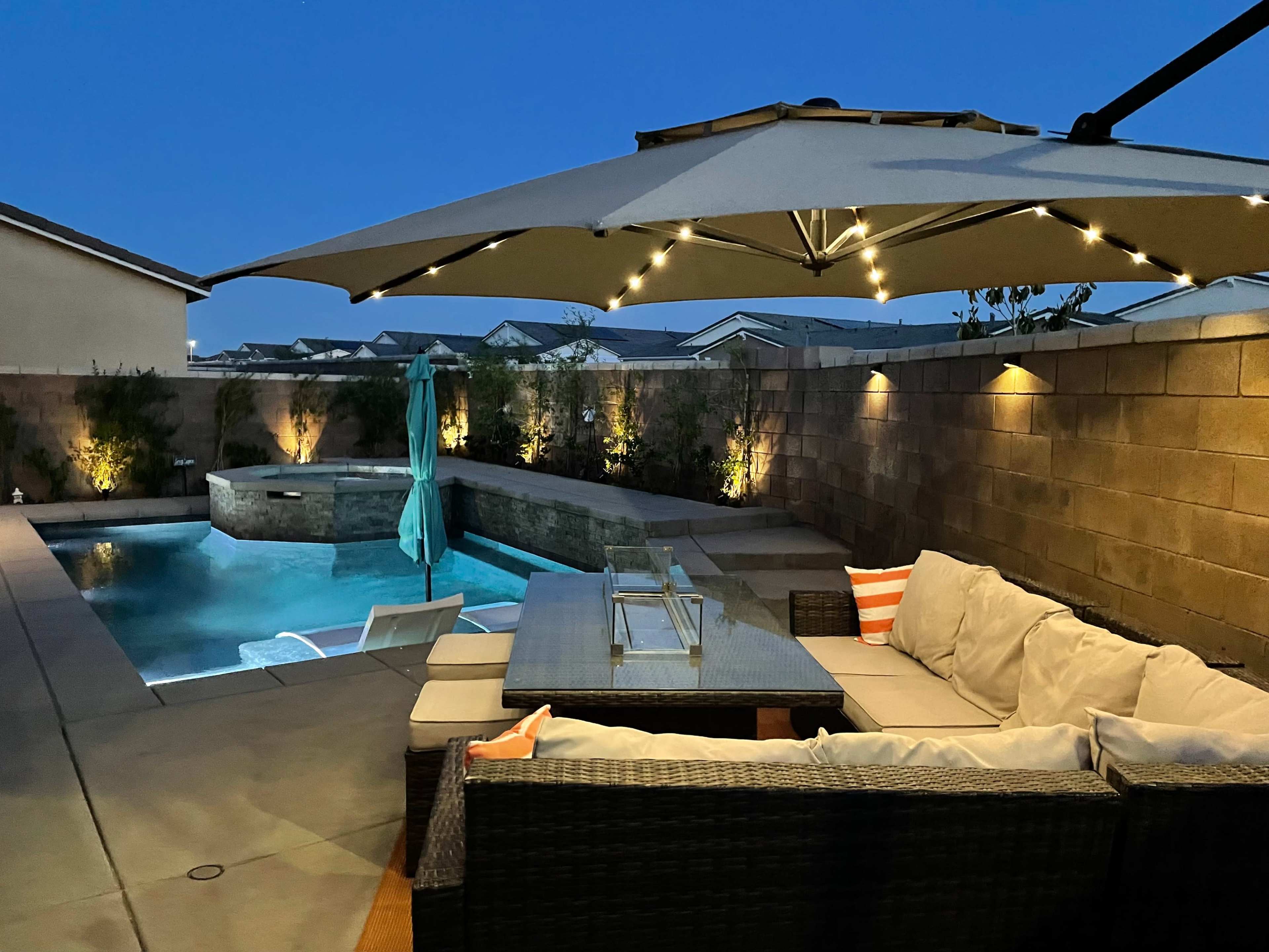 A modern outdoor lounge area features a cushioned seating arrangement beside a glowing pool, illuminated by soft lights under a large umbrella.