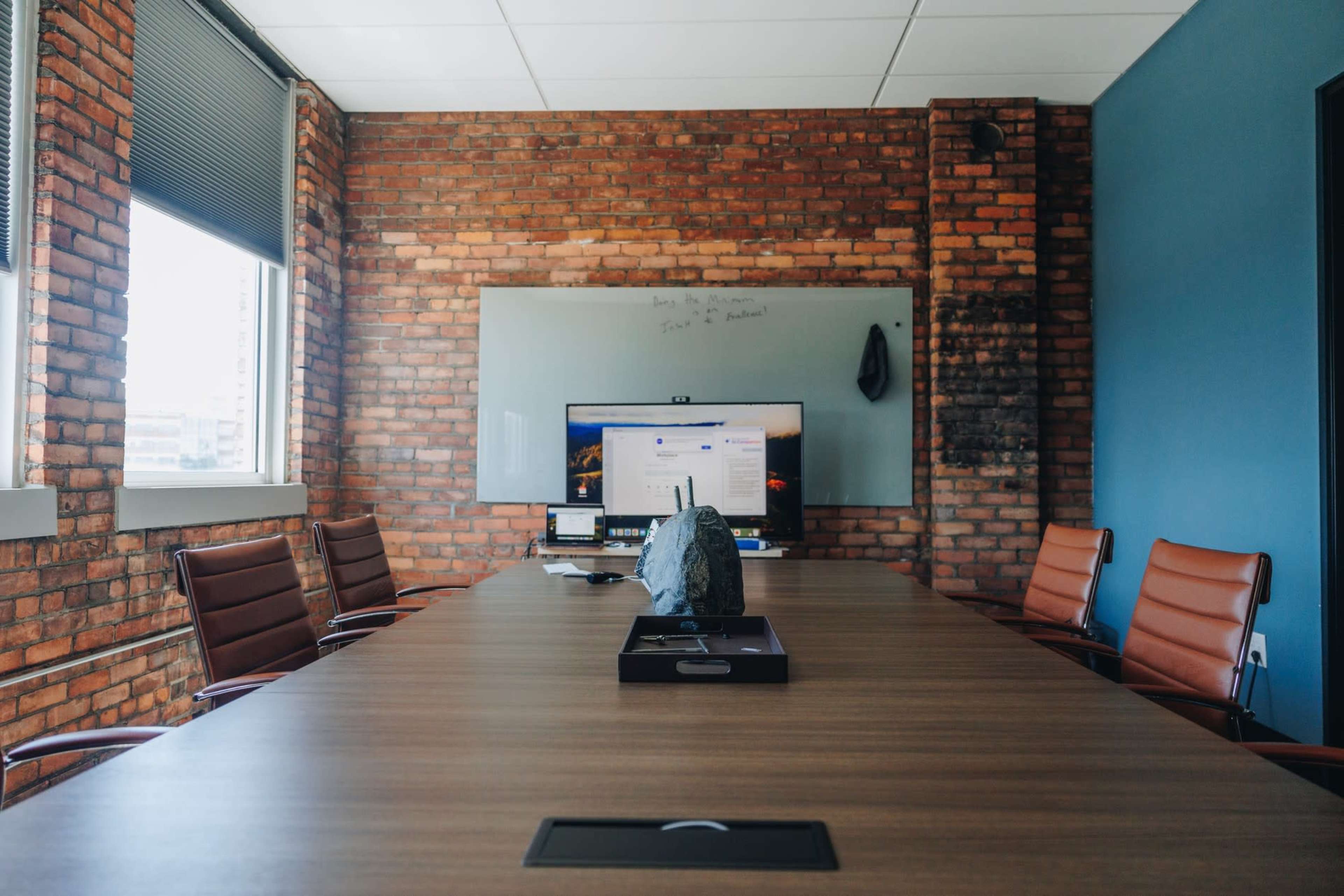 The image depicts a conference room with a long wooden table, several leather chairs, a large monitor displaying content, and a whiteboard on a brick wall.