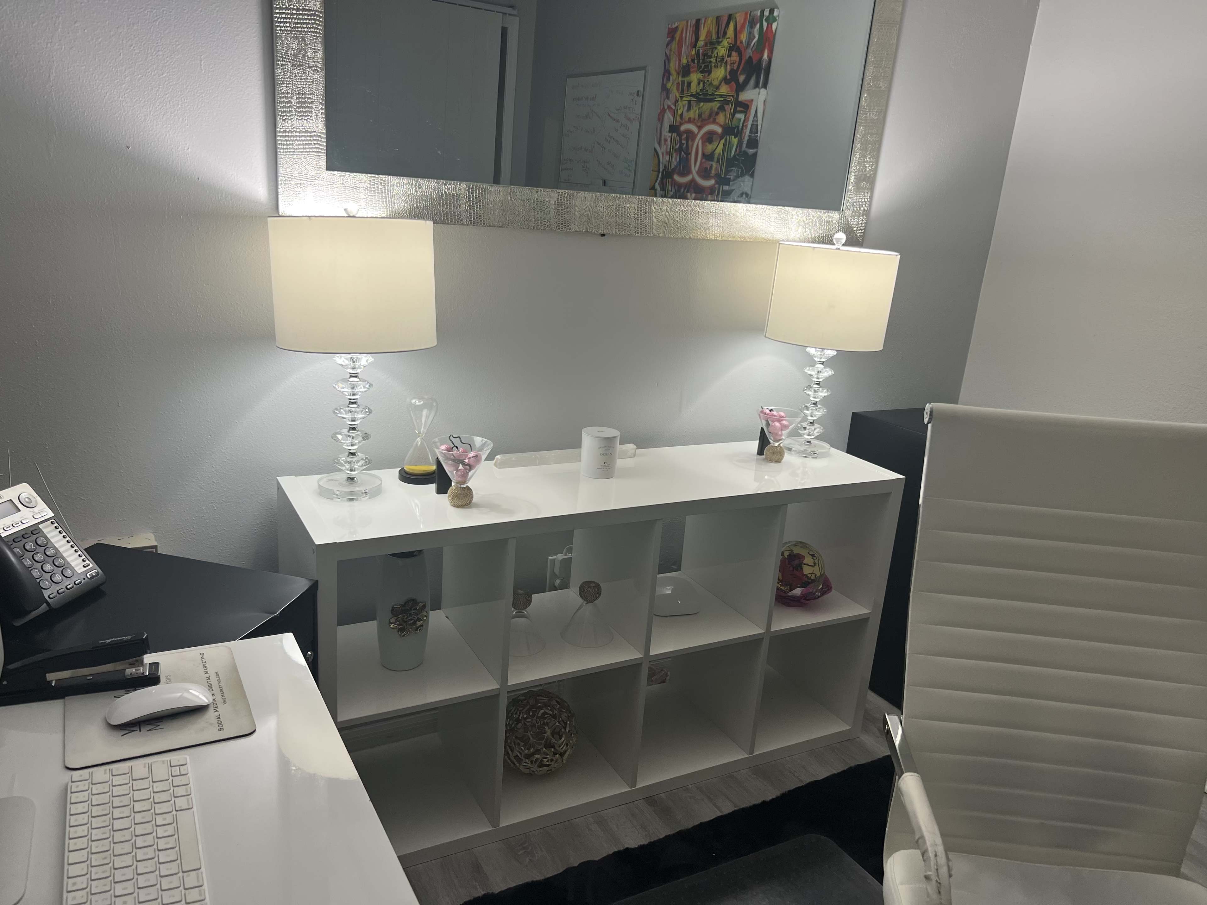A modern office space featuring a white shelving unit with decorative items, two lamps, a large mirror above, and a desk with a computer and accessories.
