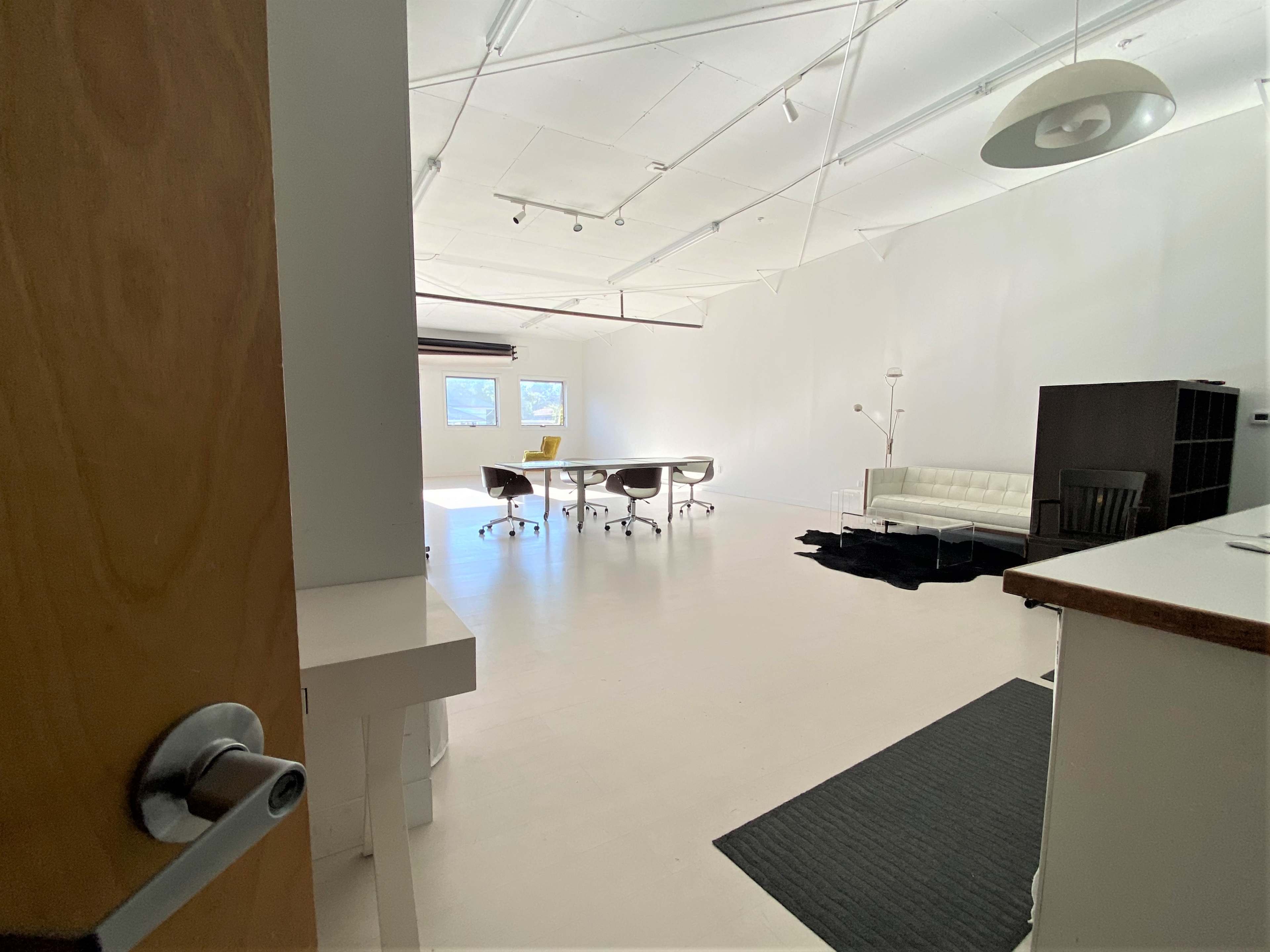 A bright, empty office space featuring a large table, several chairs, a couch, and large windows.