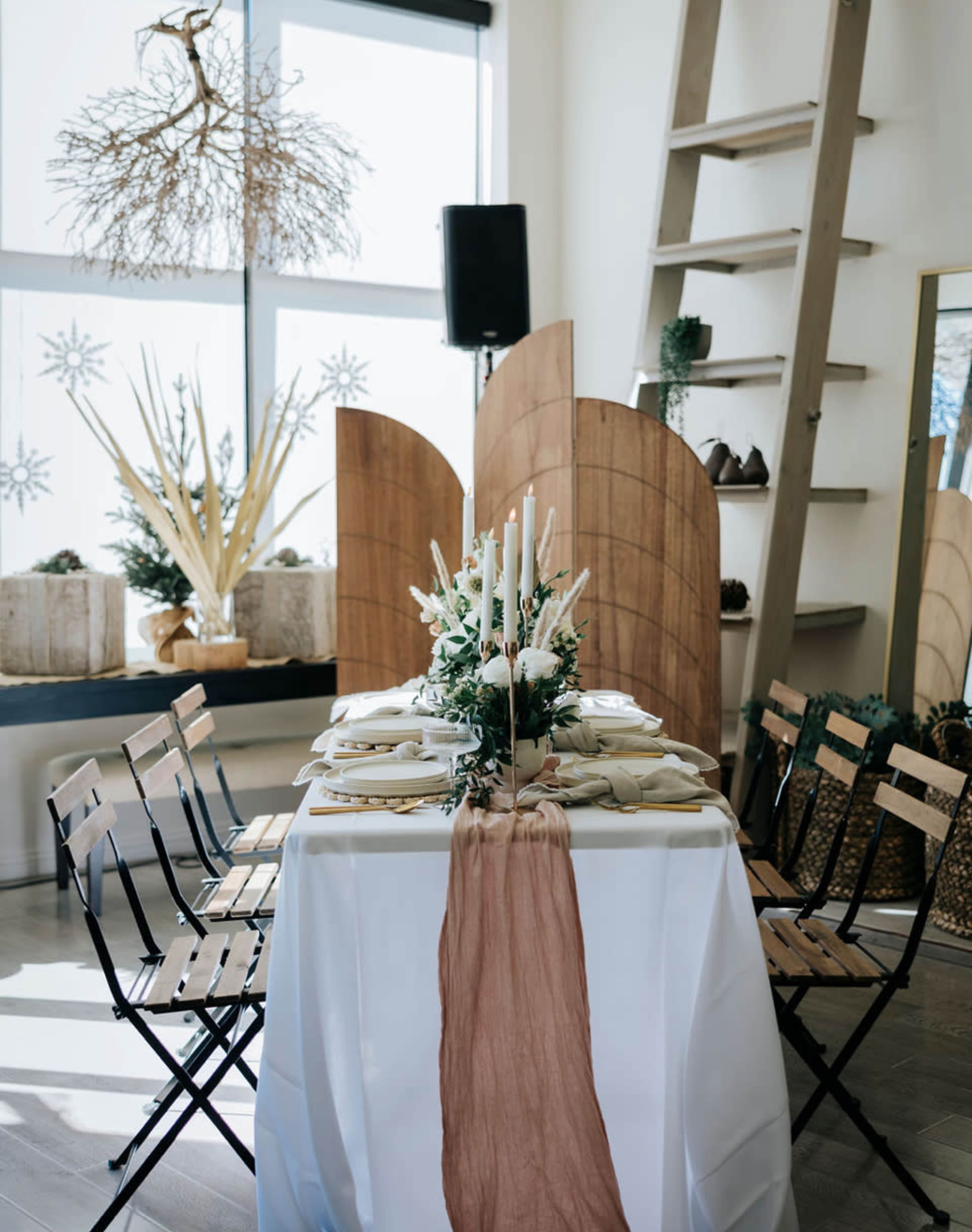 A decorated dining table with an elegant centerpiece of candles and greenery is set in a well-lit room featuring wooden accents and a ladder.