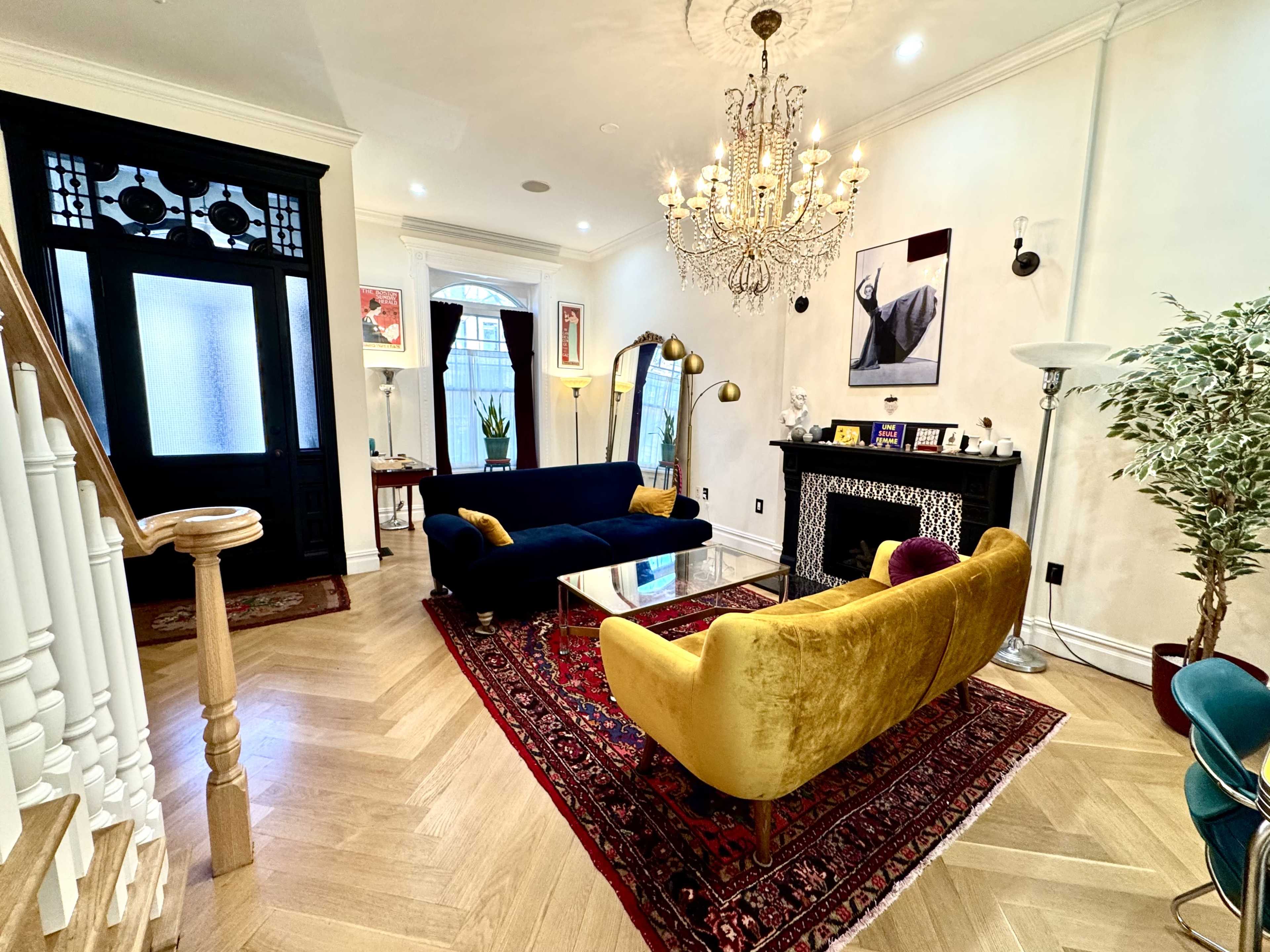 A brightly lit living room features a chandelier, a blue sofa, a yellow sofa, and a decorative fireplace, with a patterned rug and various artworks on the walls.