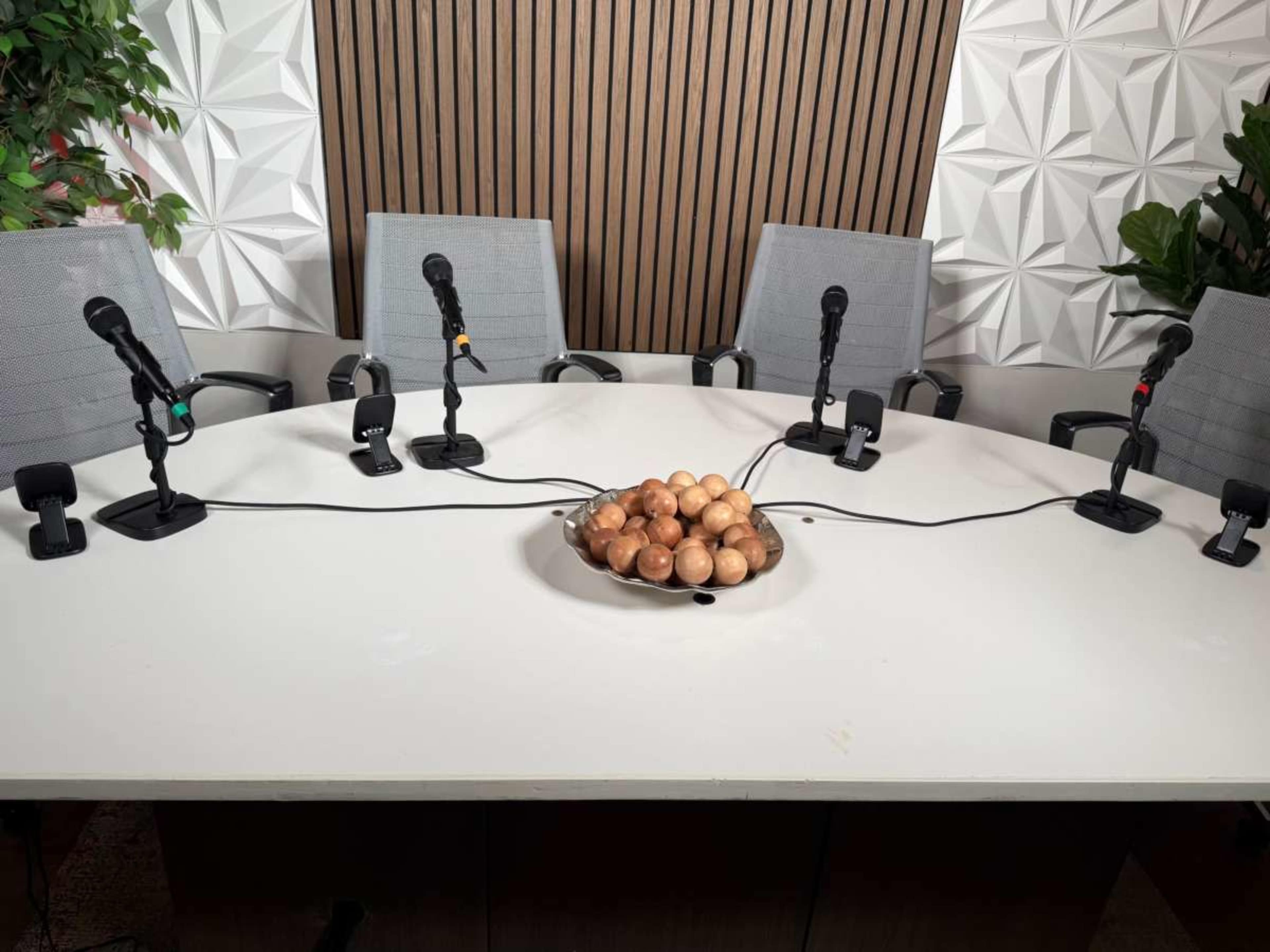A round table is set with several microphones and a bowl of brown wooden balls at the center, surrounded by four gray chairs.