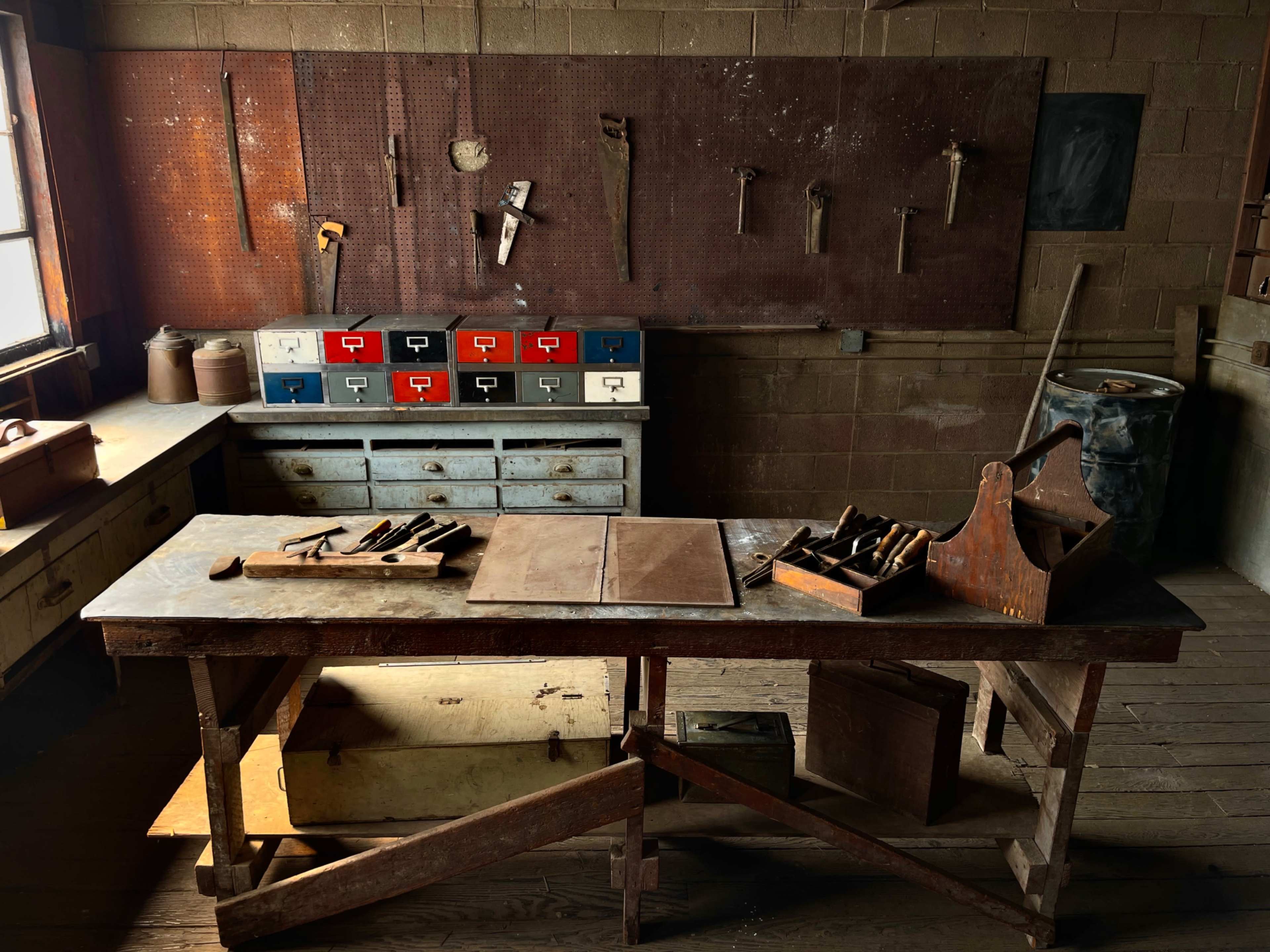 A wooden workbench with various tools and a collection of drawers sits in a dimly lit workshop with a tool-laden wall behind it.