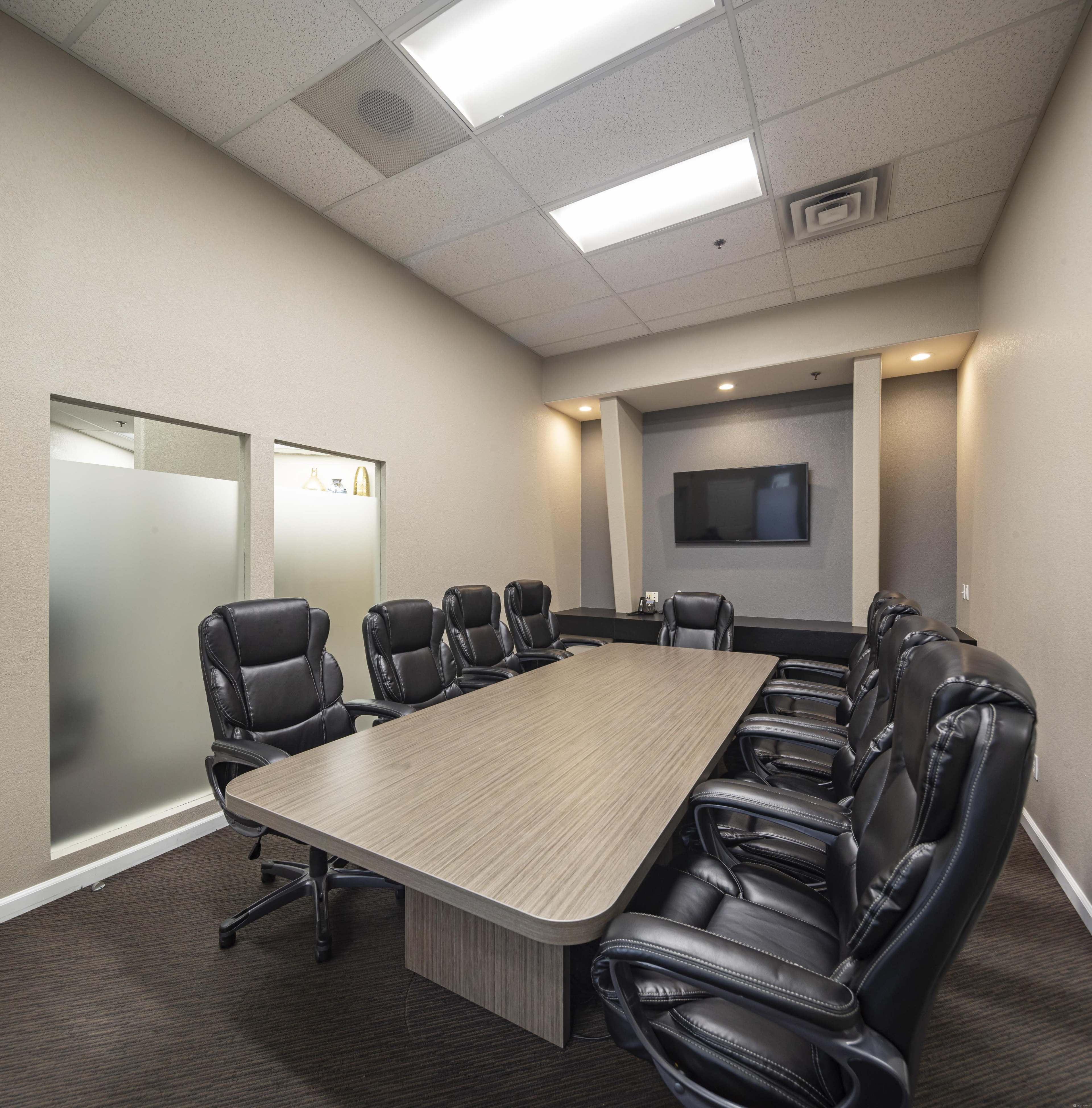 A modern conference room features a long wooden table surrounded by black leather chairs and a wall-mounted TV.