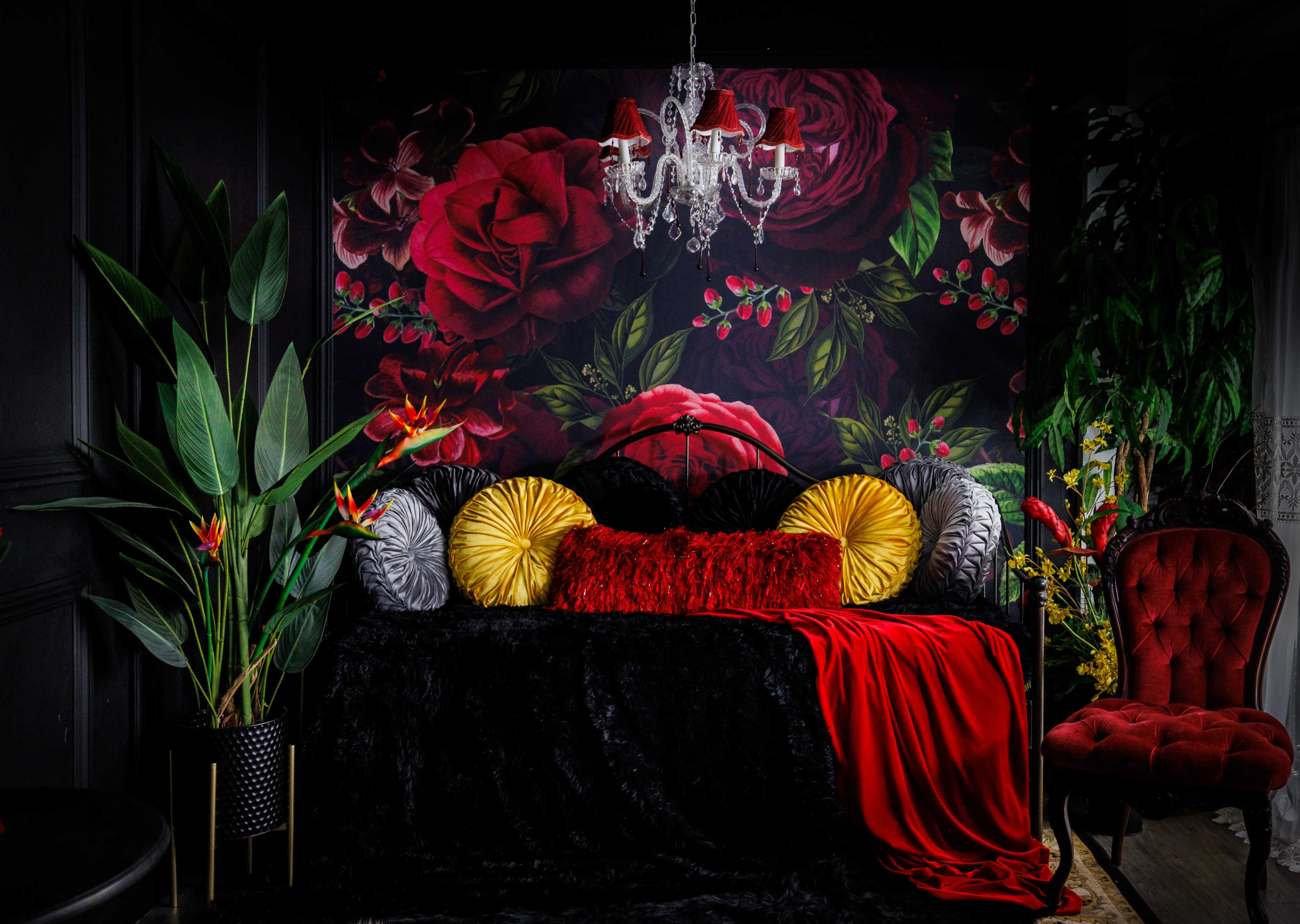 The image features a dark room with a floral wallpaper mural, a black sofa adorned with colorful cushions and a red throw, a chandelier above, and a large decorative plant.