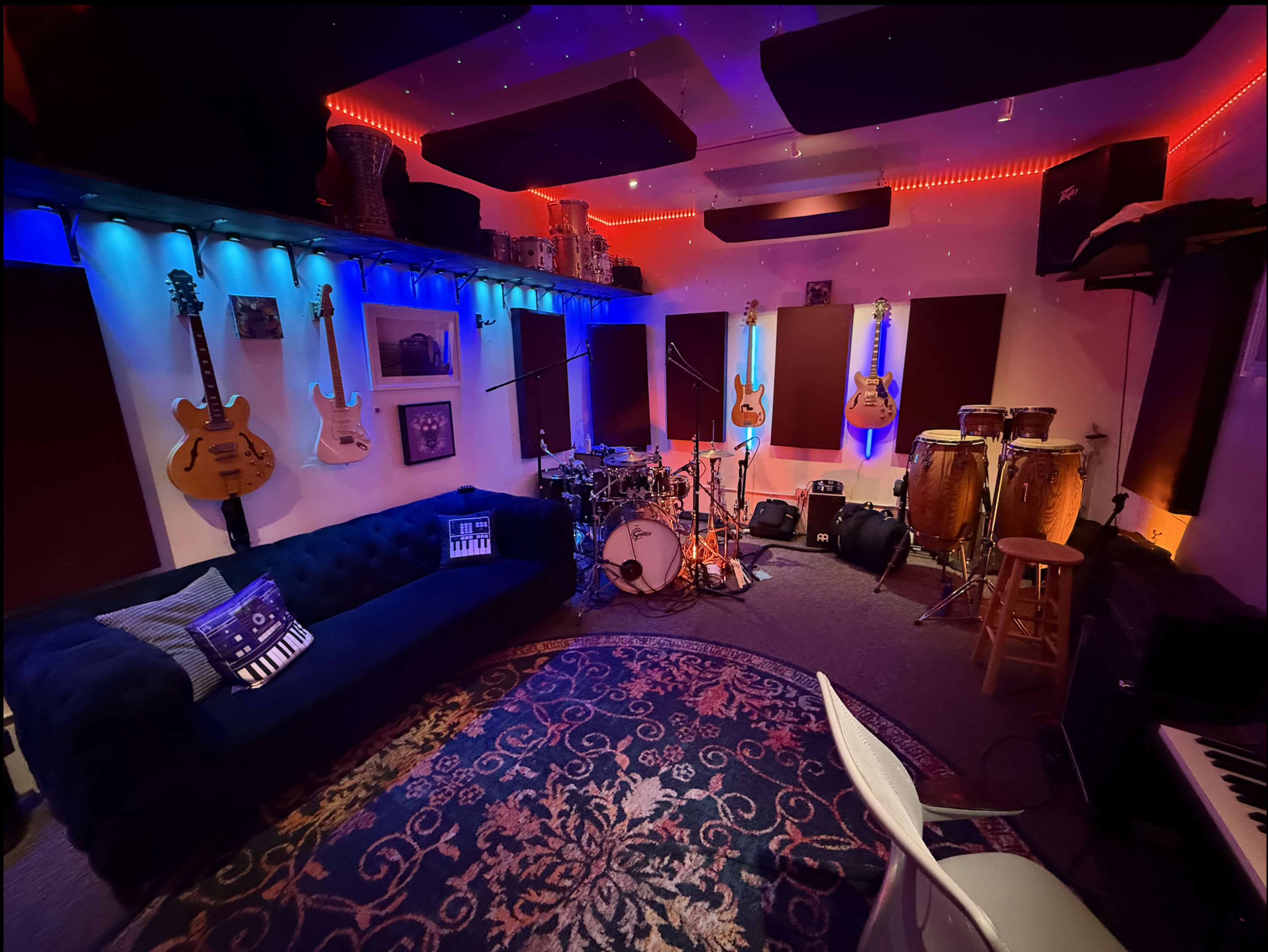The image shows a vibrant music studio with bright lighting, featuring guitars mounted on the walls, a drum set, conga drums, and a comfortable couch on a patterned rug.