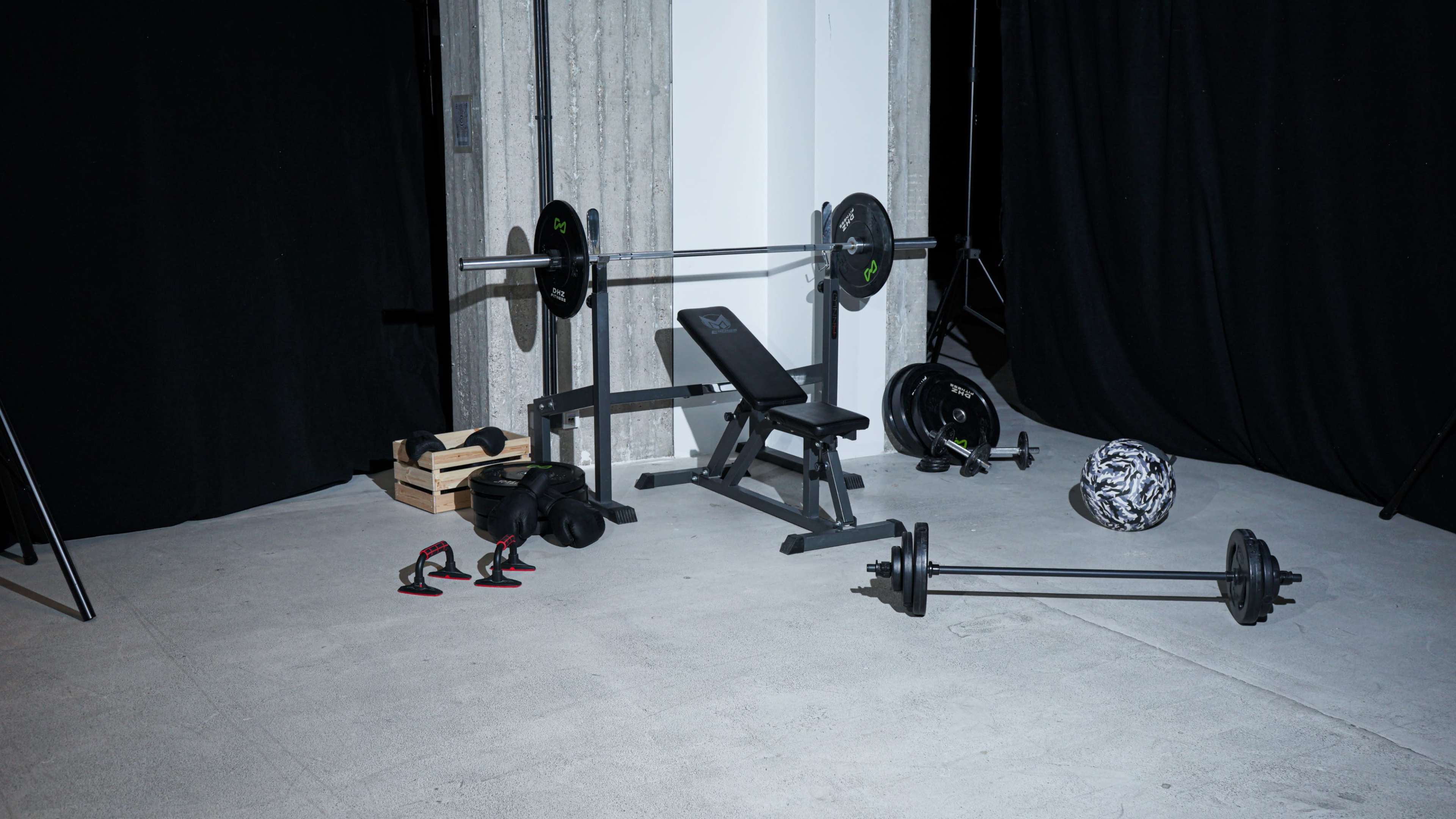 The image shows a gym setup with a weight bench, barbell, weights, resistance bands, and exercise equipment arranged on a concrete floor.