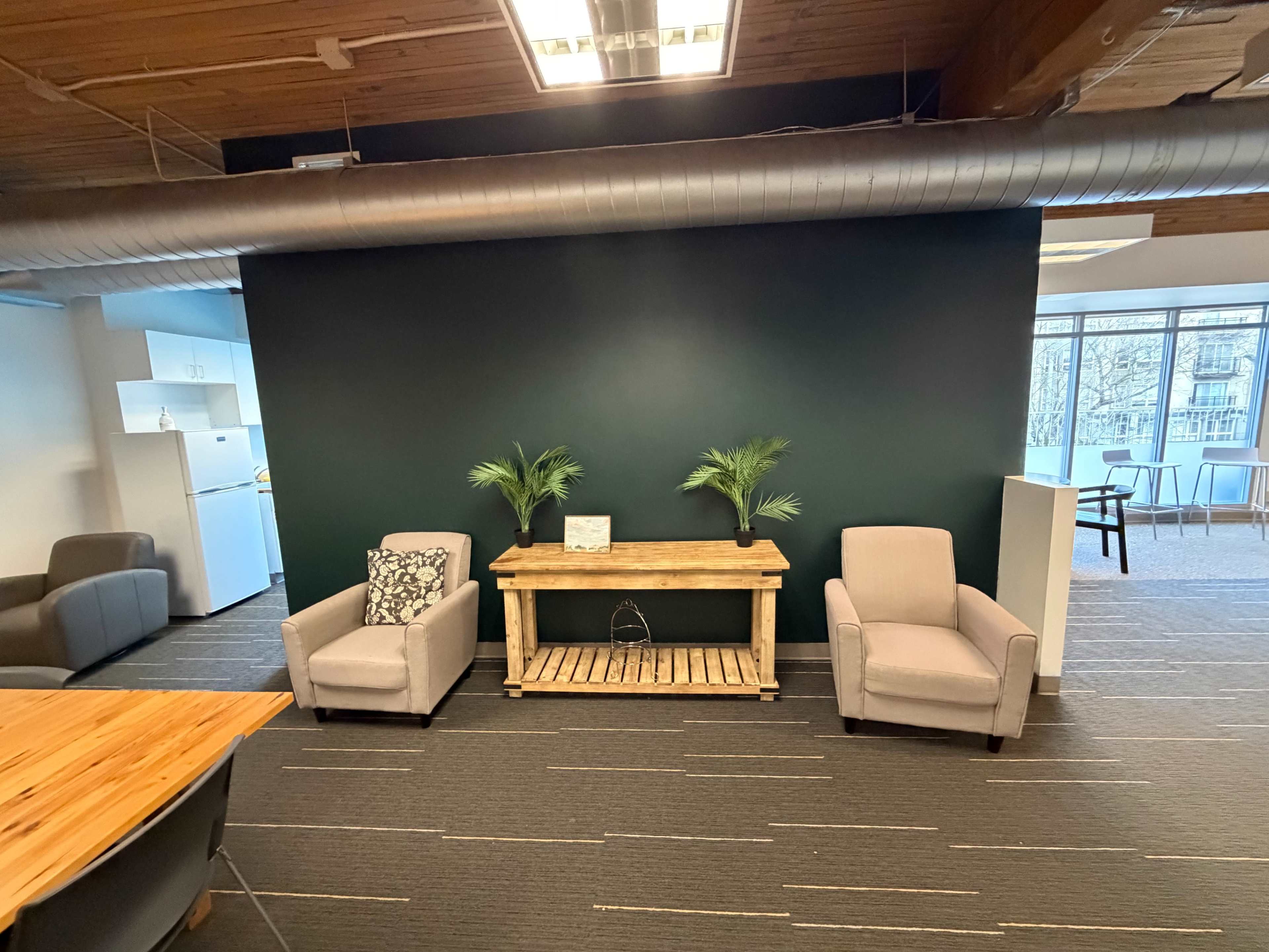 The image shows a modern lounge area featuring two light-colored chairs, a wooden table with decorative plants, and a dark green accent wall.