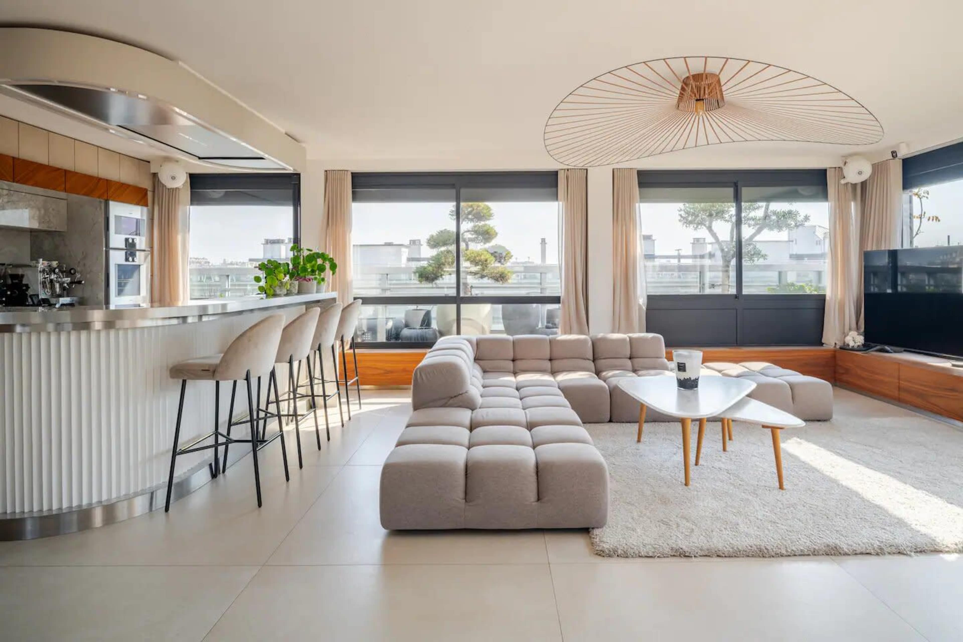 A modern living room with a large, light-colored sectional sofa, a coffee table, and a kitchen area featuring bar stools, all illuminated by natural light from large windows.